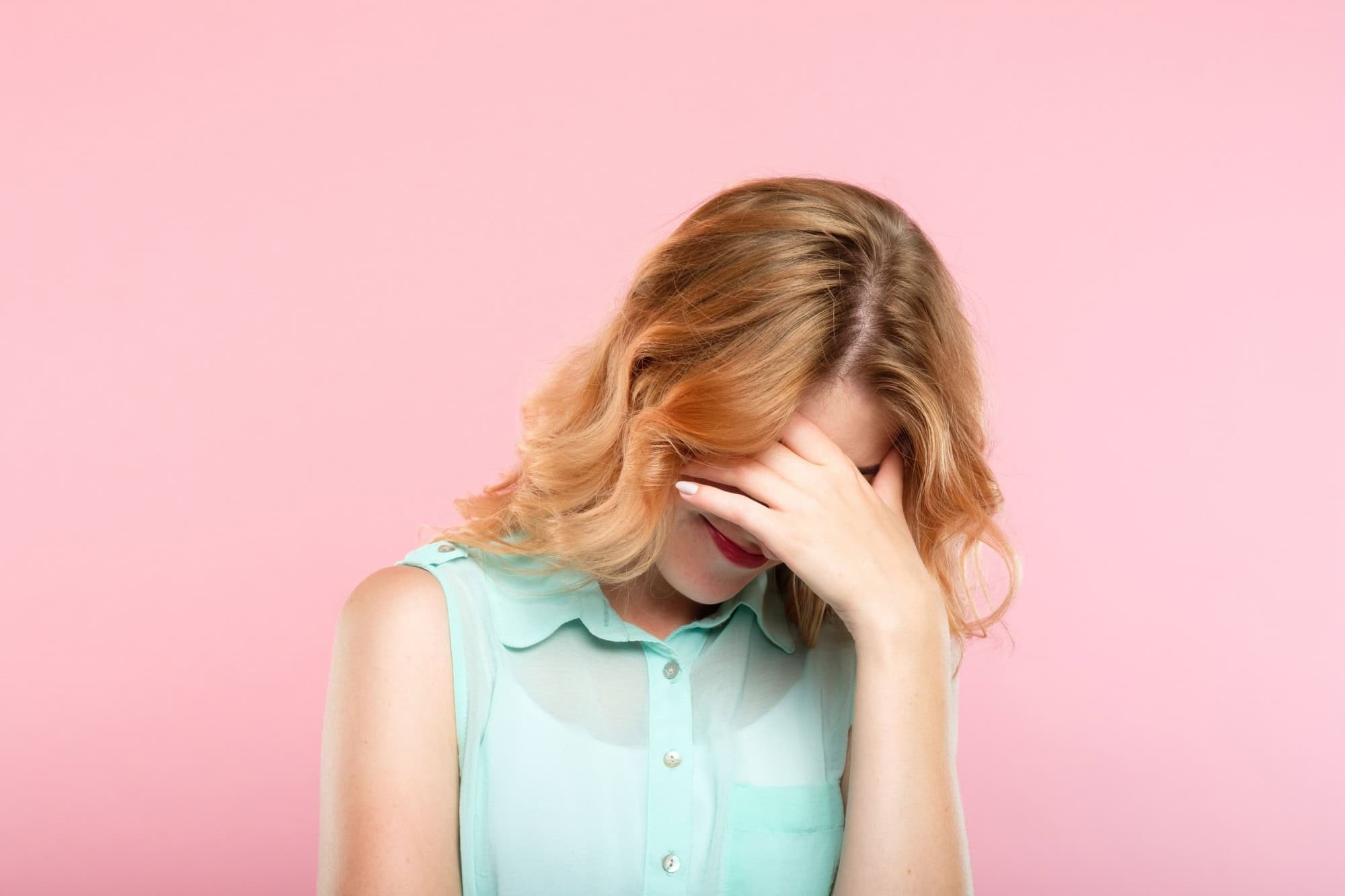 facepalm embarrassment and shame emotion. ashamed smiling girl covering her face with a hand. young beautiful woman portrait on pink background.