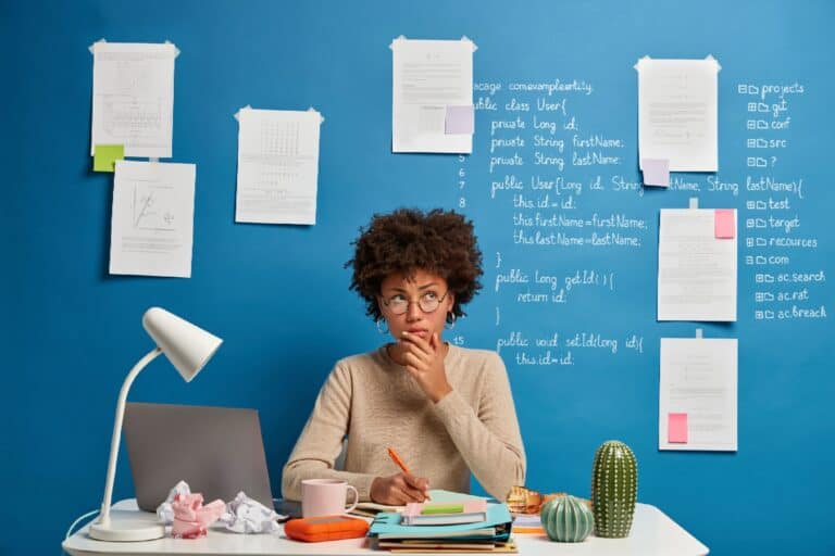 Thoughtful attractive woman in spectacles writes in diary to do list of goals, makes notes in personal organizer, poses at cozy coworking space, creats article on scientific topic, thinks of plans