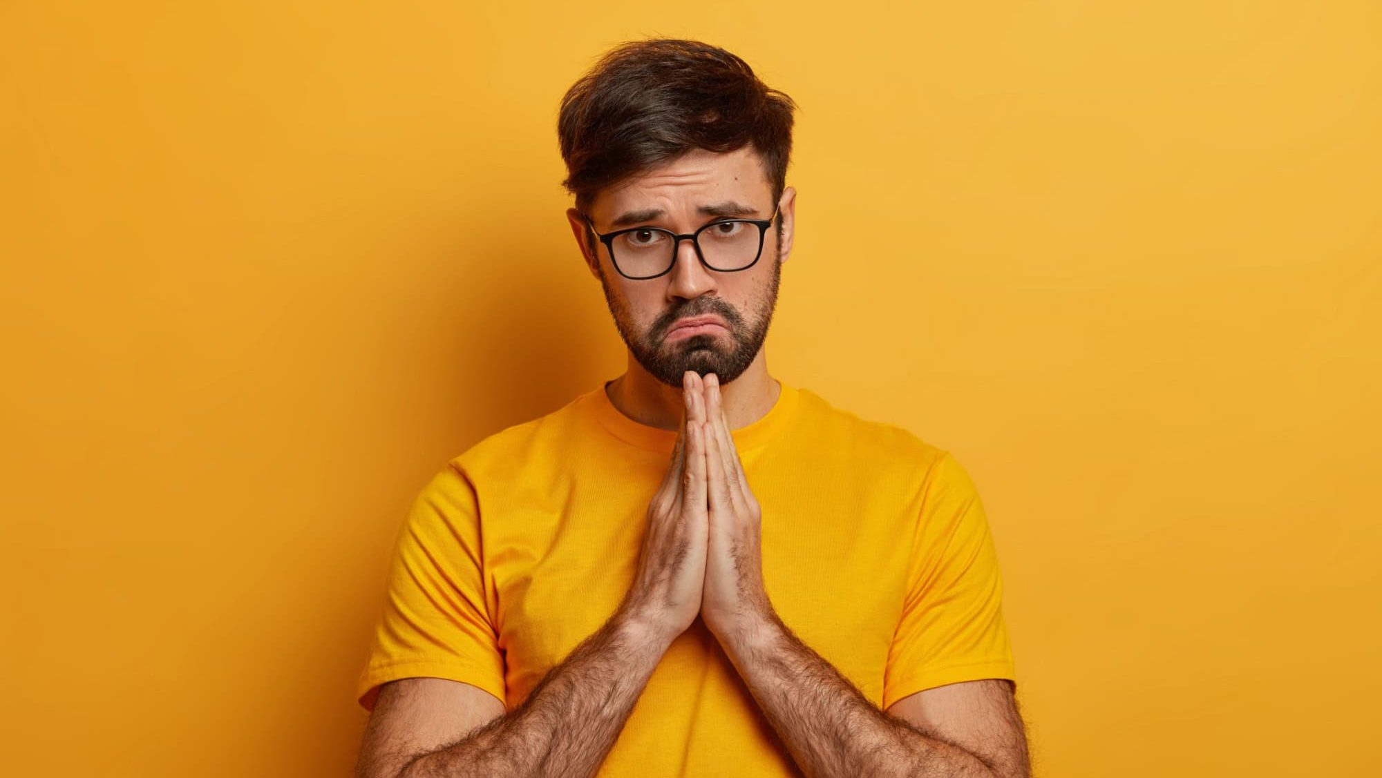 Miserable sad pleading man says sincere please, makes apology, keeps palms pressed together, begs with upset expression, needs help in trouble, prays with hope has faith in better wears yellow t shirt