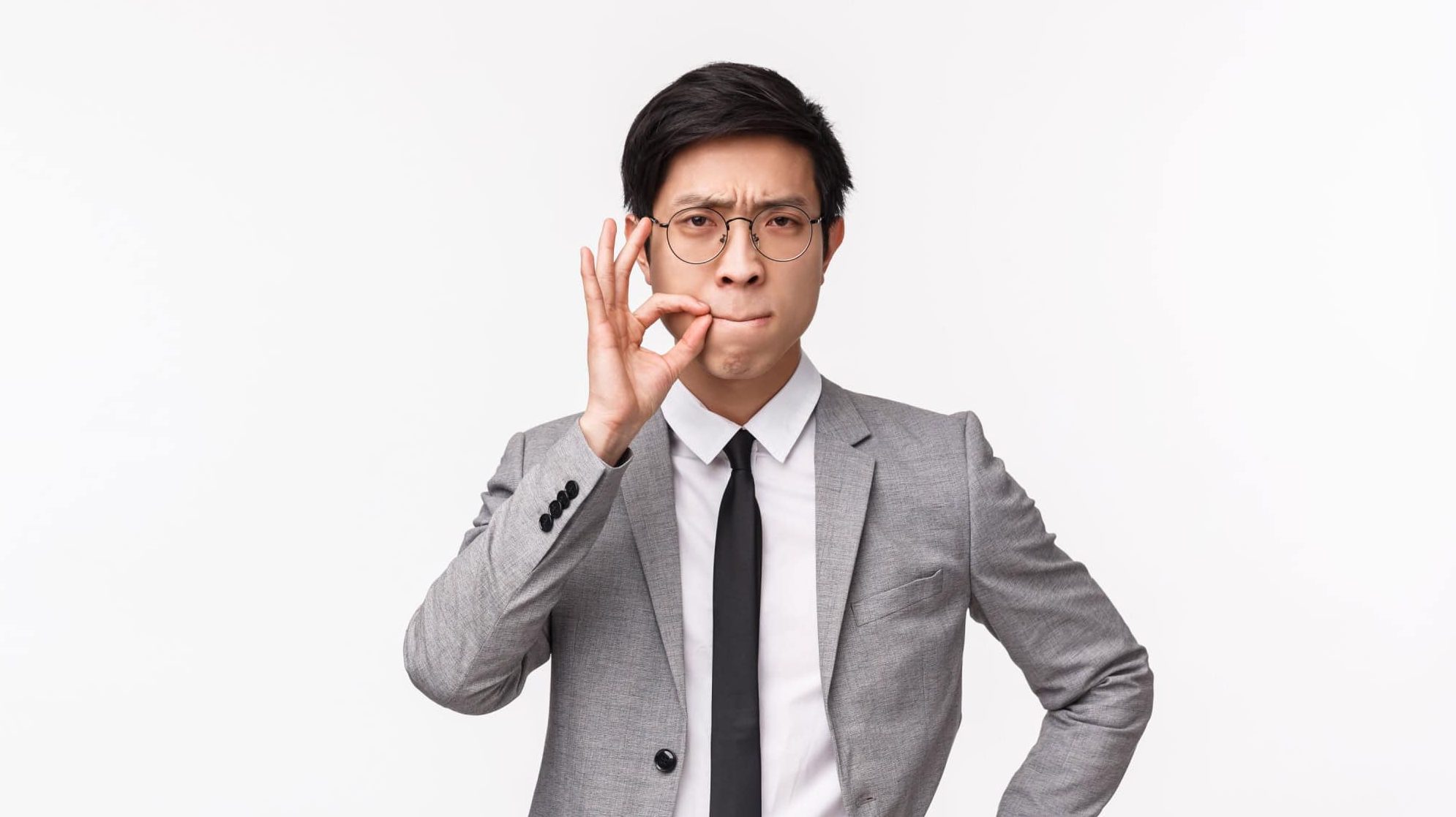 Keep silent about this conversation. Serious-looking asian guy in grey suit, zipping his lips on lock, promise not tell, hiding secret, asking be quiet, standing white background