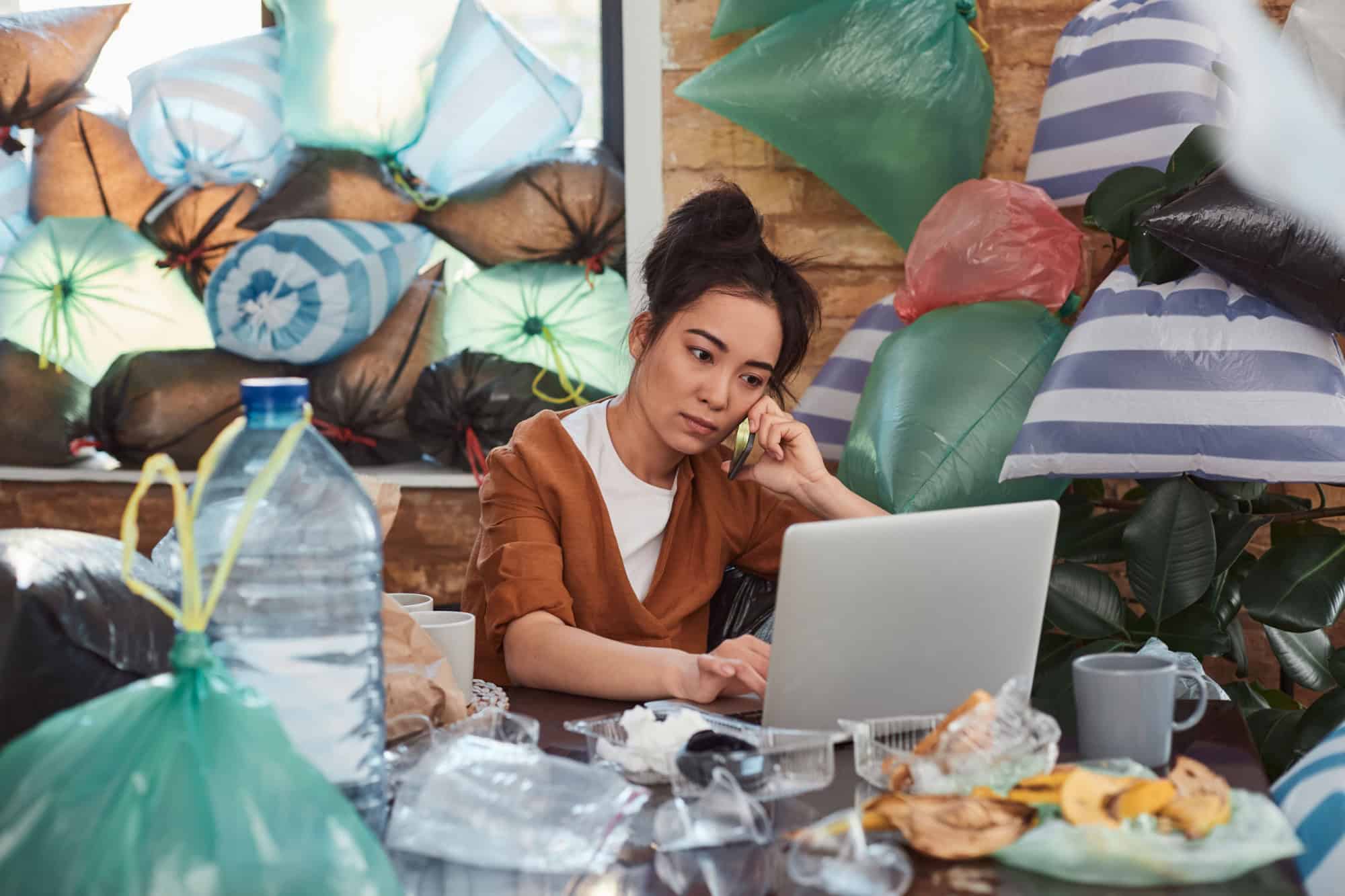 Slatternly Asian woman sitting before laptop and holding smartphone close to ear among leftovers, plastic bags and rubbish on background