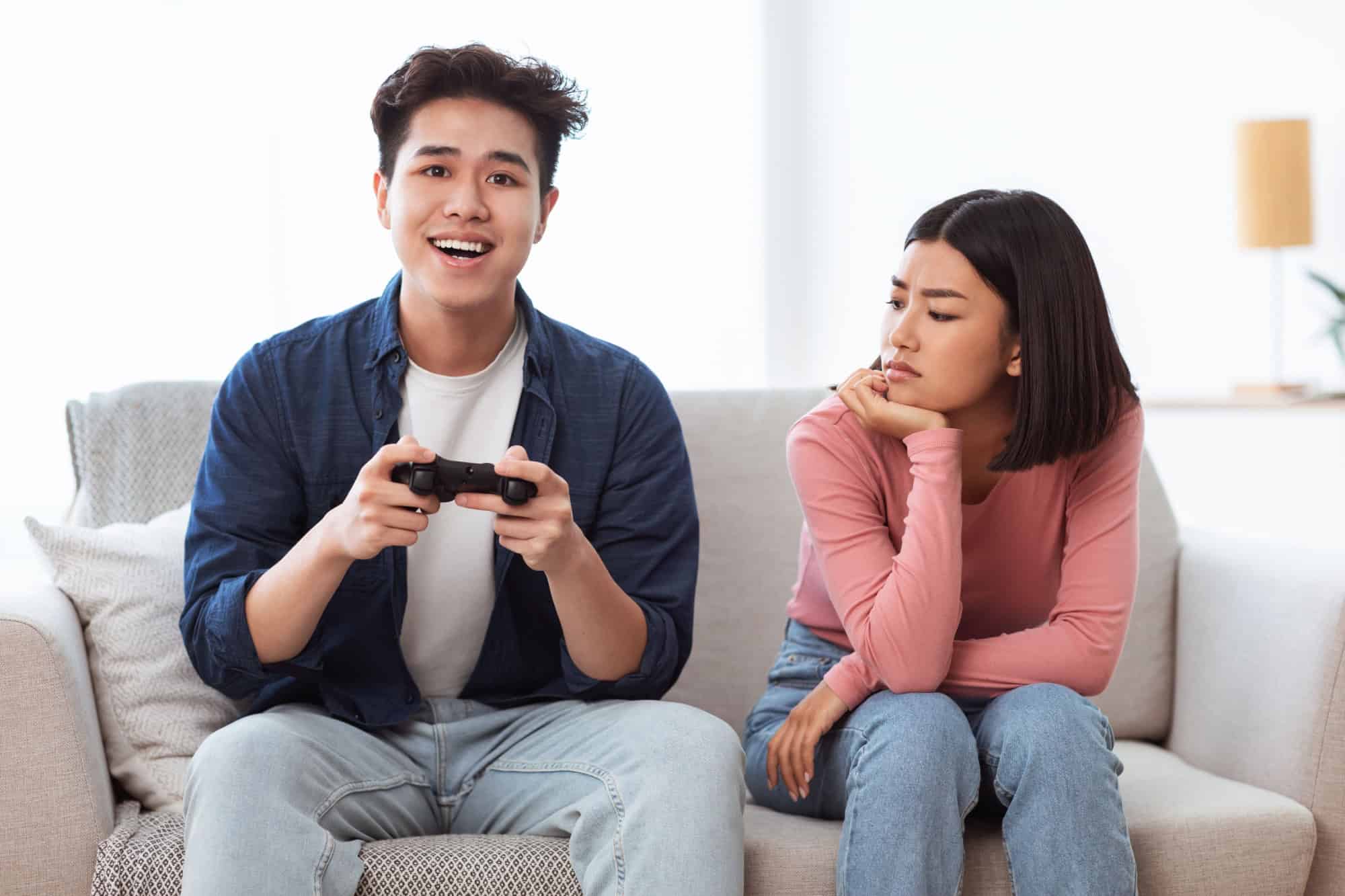 Gaming Addiction Problem. Discontented Asian Wife Looking At Infantile Husband Playing Video Game Sitting On Couch At Home. Computer Games, Waste Of Time, Male Emotional Immaturity Concept