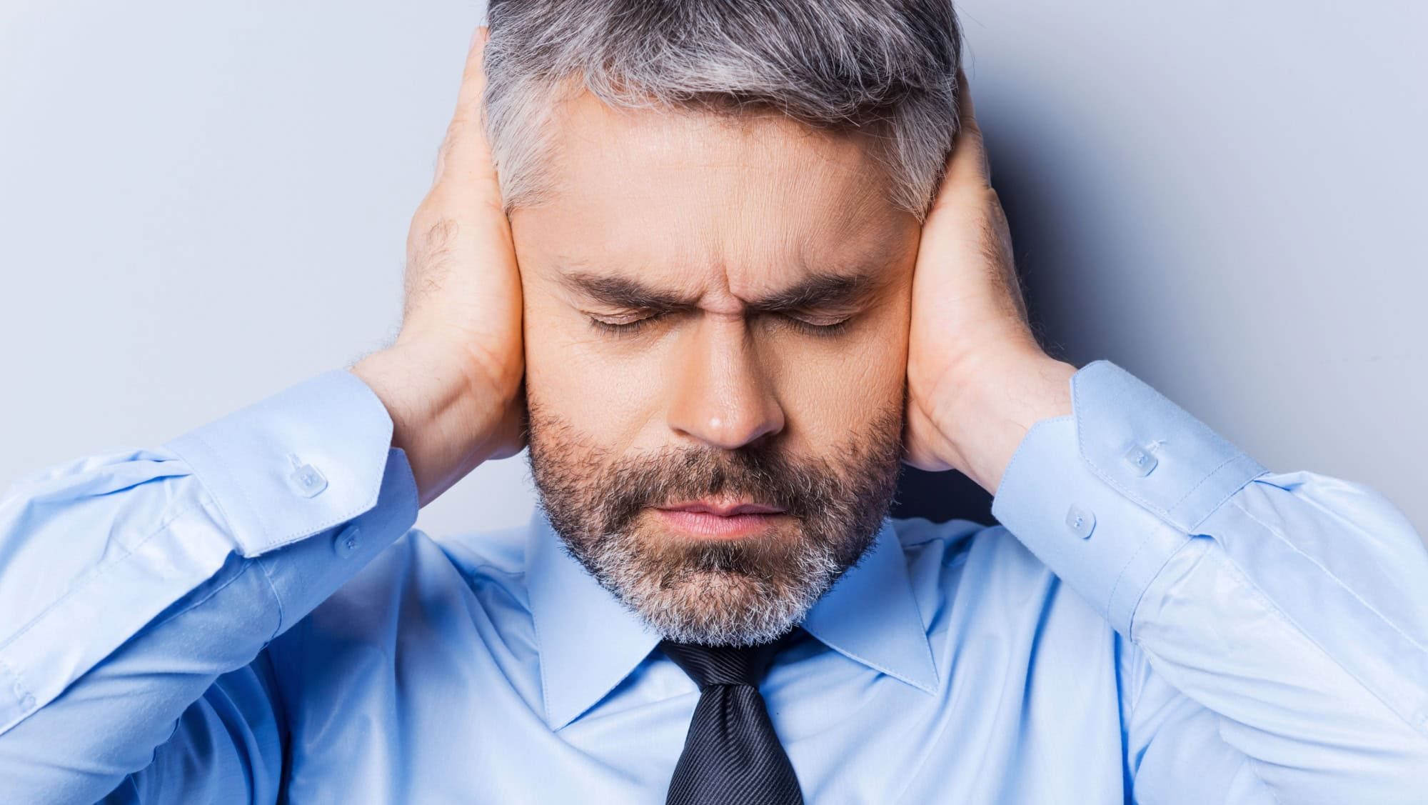 Feeling stressed and overworked. Frustrated mature man in shirt and tie covering ears with hands and keeping eyes closed while standing against grey background