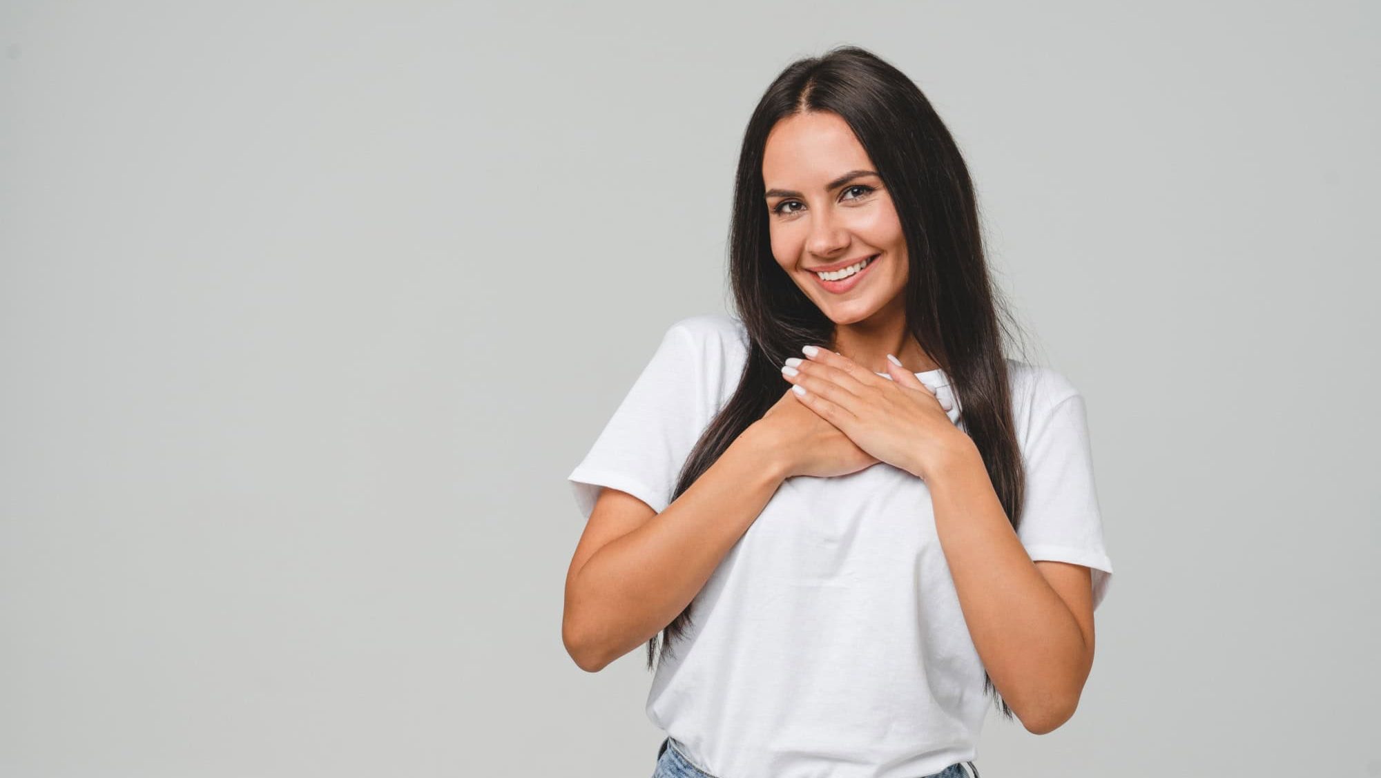 Touched pleased with a compliment cheerful happy caucasian young woman in white T-shirt isolated in grey background. Praise recommendation, sweet heart