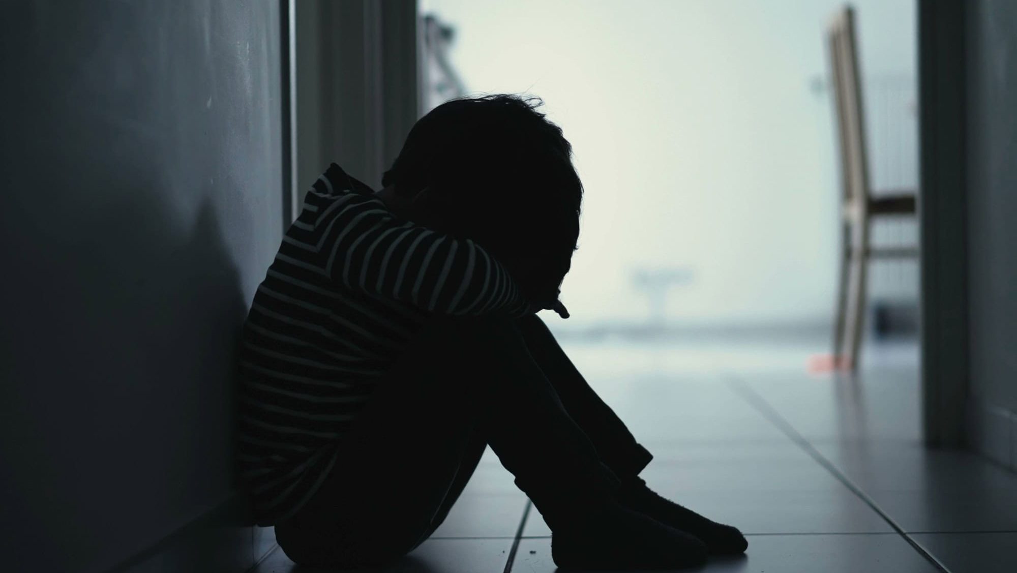 Sad child suffering from depression sitting alone in corridor feeling loneliness. Scared fearful small boy covering face in silhouette at home