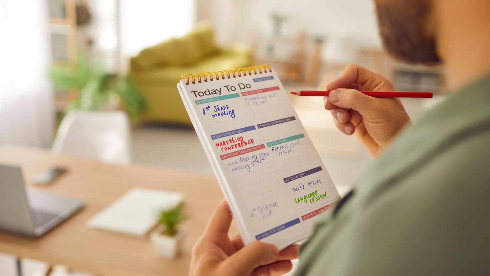 Close up photo of business man holding to do list in hands writing in his notepad standing at home or at office. Male person planning daily appointment and tasks in planner schedule.