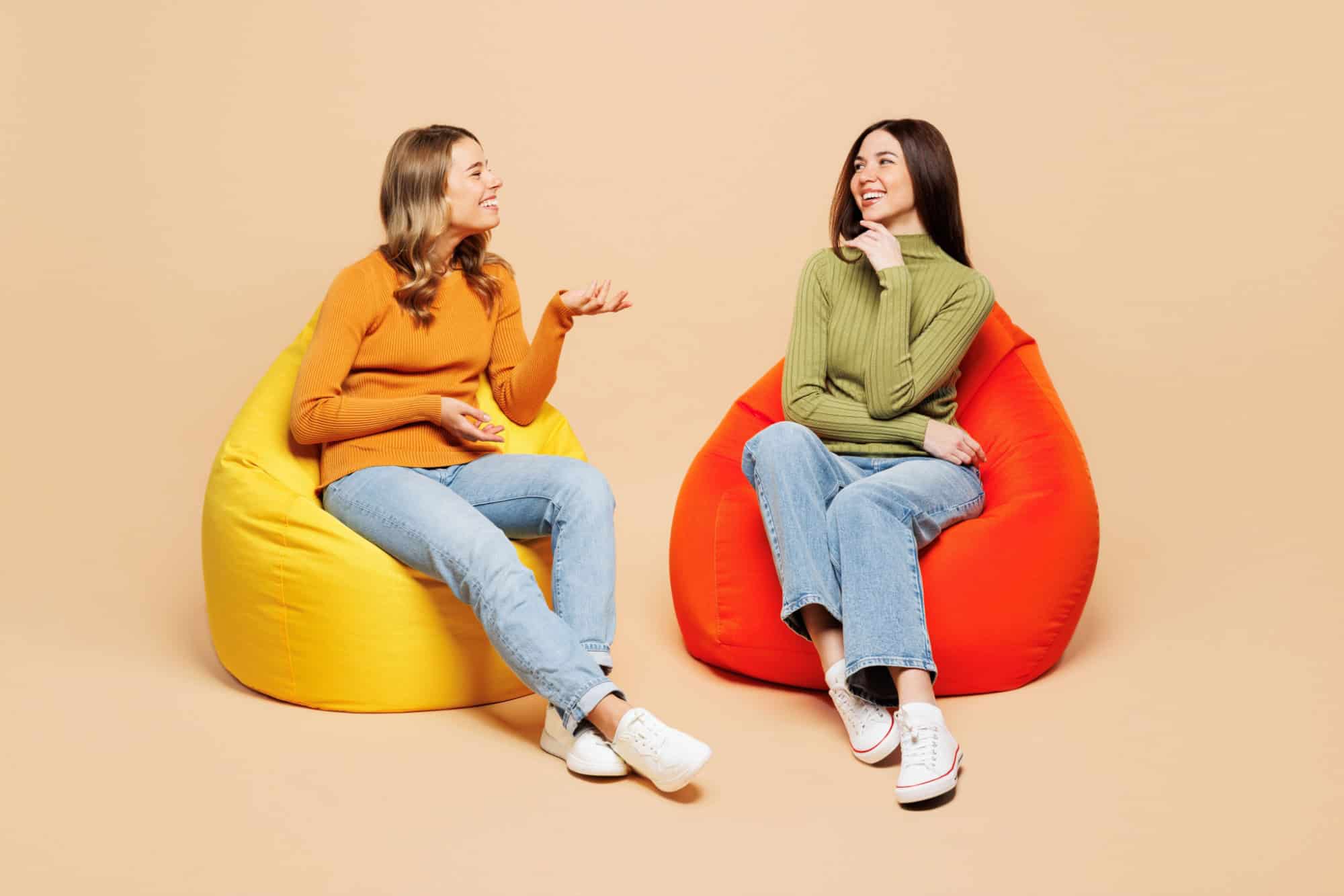 Full body young friends two women they wear orange green shirt casual clothes together sit in bag chair talk with each other isolated on plain pastel light beige background studio. Lifestyle concept