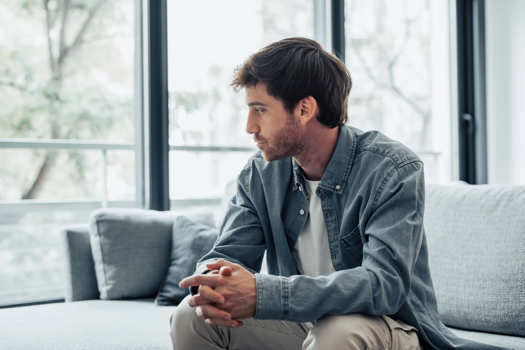 Thoughtful serious man sitting on sofa alone at home, lost in thoughts, thinking about problem solving, feeling lonely, making important decision, having psychological problem, side view