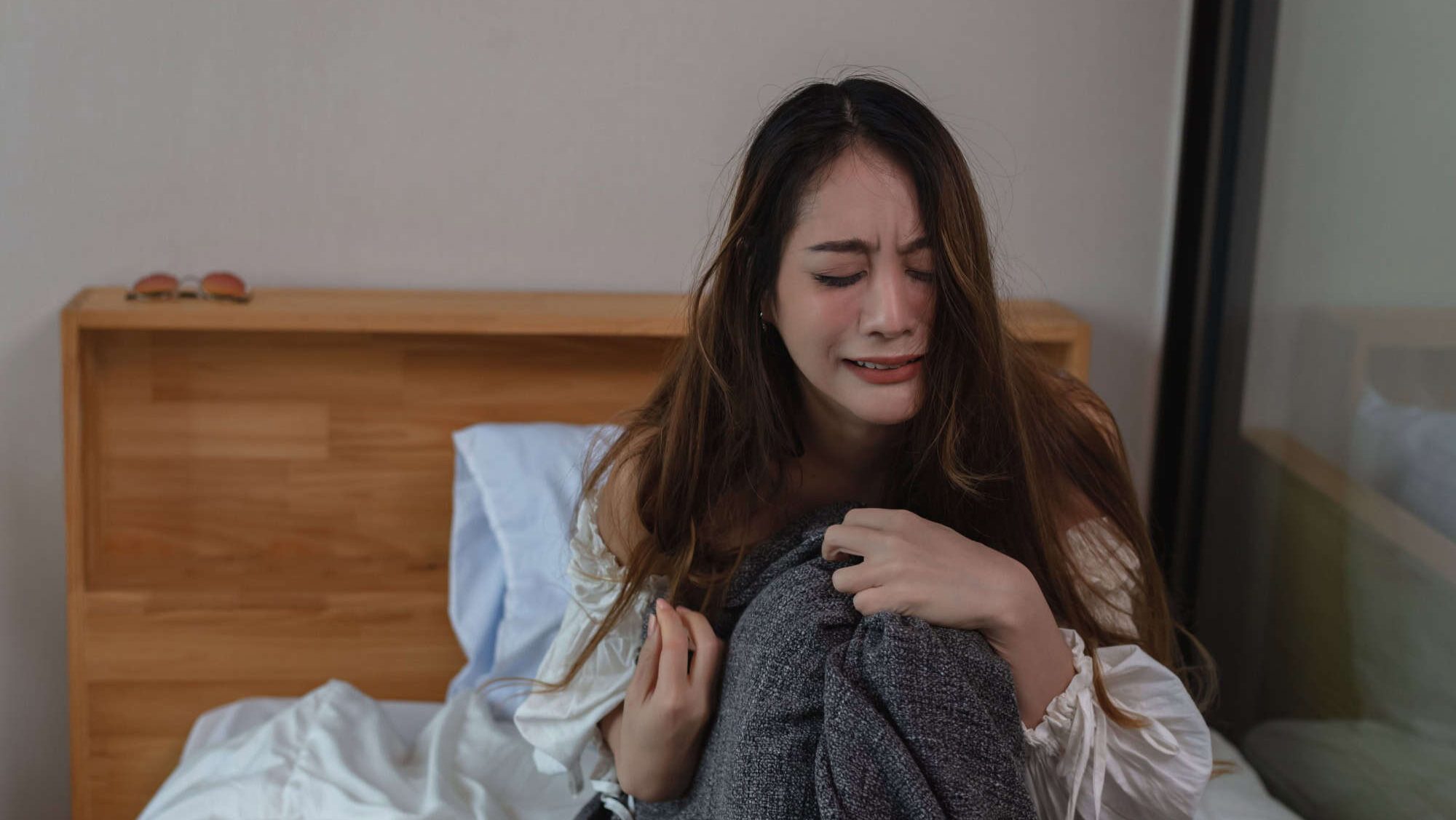 Woman sitting alone on the bed in dark bedroom with hugging self and crying. Loneliness sad girl thinking grief trouble life problems.