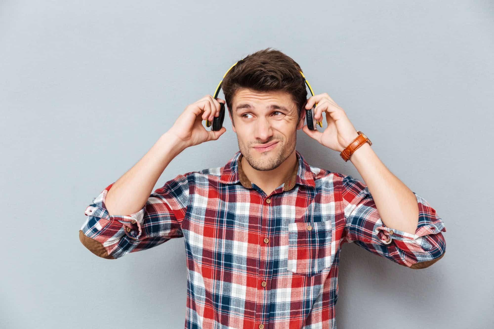 Confused unhappy young man in headphones listening to music over grey background