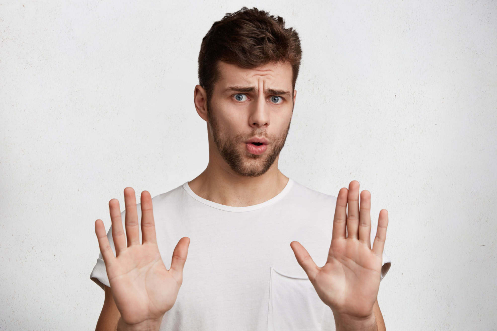 Attractive brunet male shows refusal gesture, doesn`t want to participate in meeting, says: It`s not for me, leave me in piece, has angry expression, poses against white concrete studio background