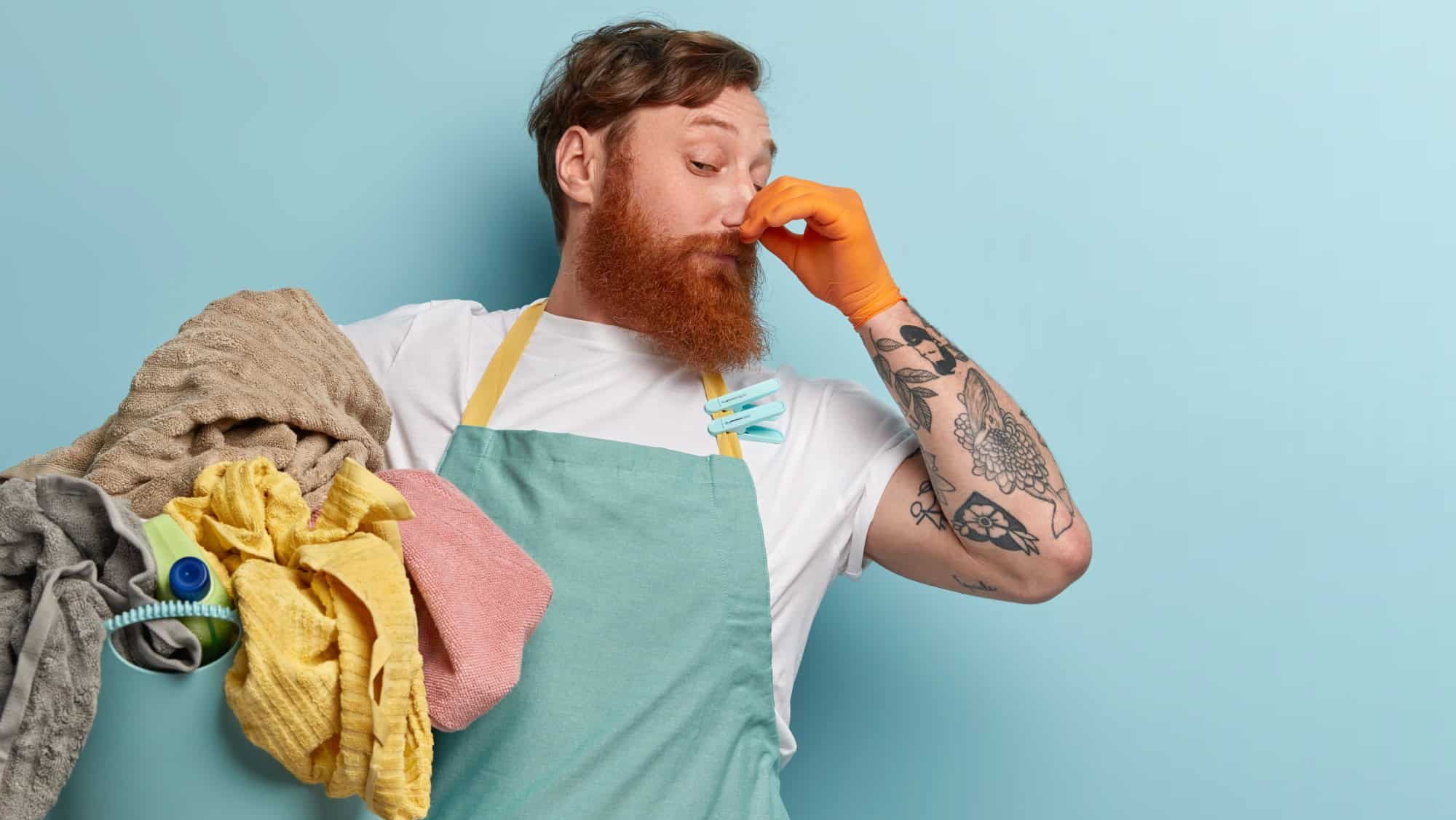 Foxy bearded man closes nose with fingers from unpleasant smell, collects all dirty laundry, wears casual t shirt and apron with clothespins, has tattoo, stands over blue background, free space