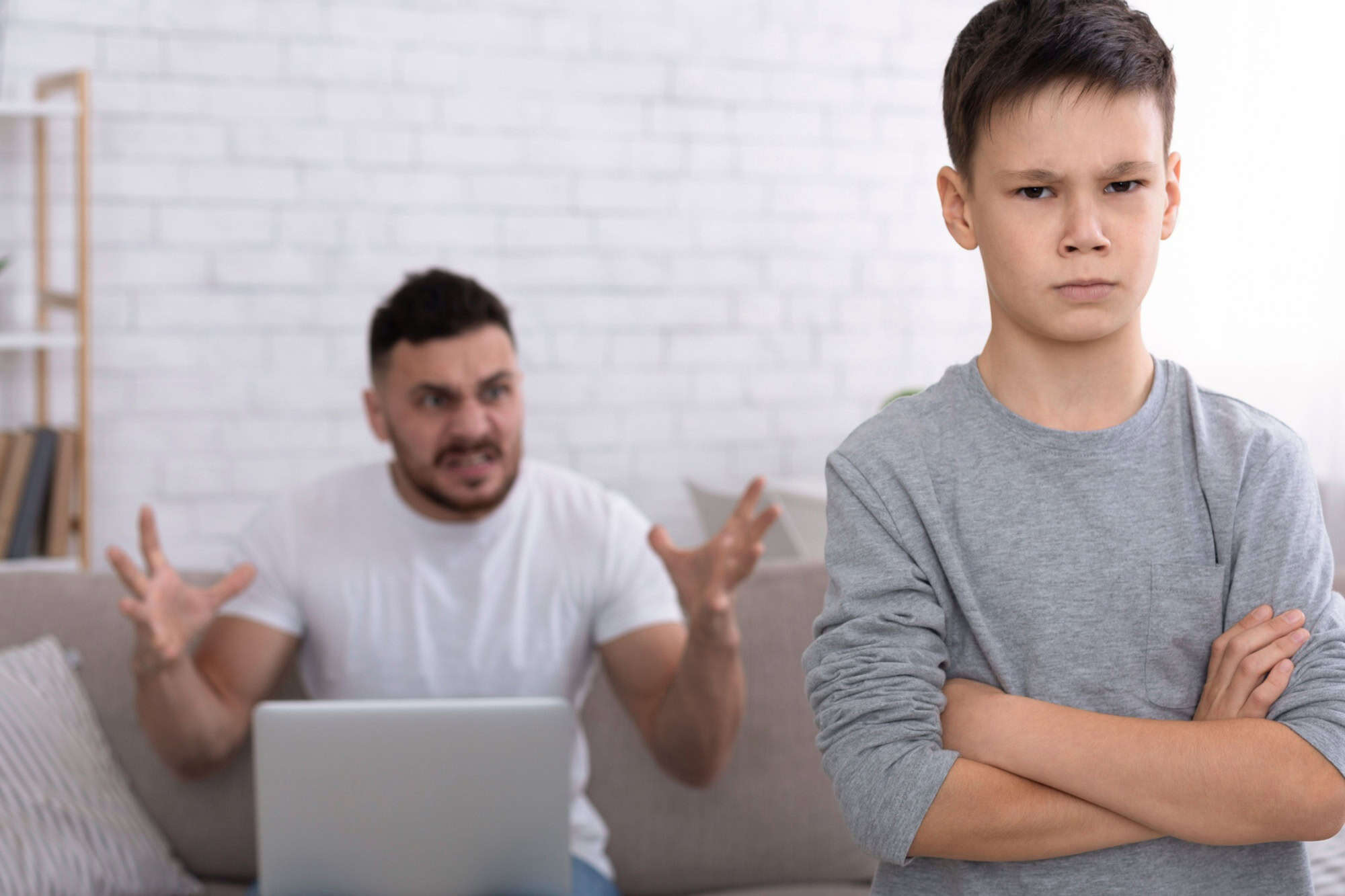 Generations misunderstanding. Sad little boy looking at camera with crossed hands, angry father with laptop yelling at him in background, free space