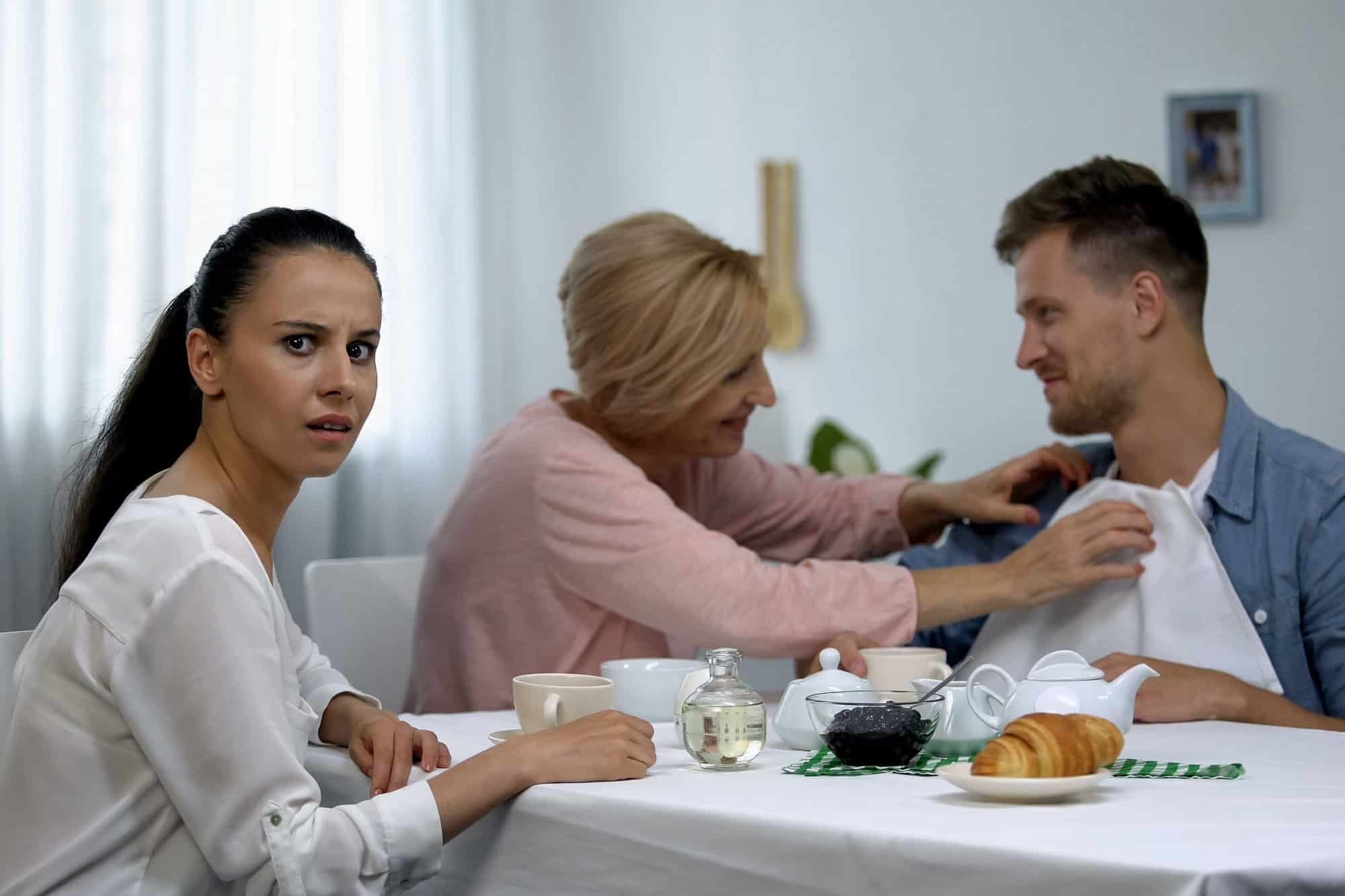 Shocked wife looking at mother-in-law putting napkin on husbands neck, tea party