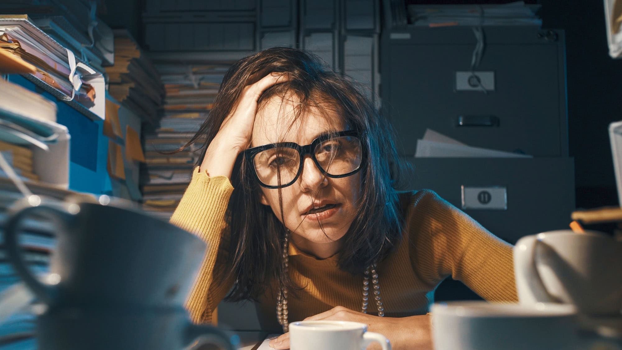 Stressed exhausted woman sitting at office desk and working overtime, she is overloaded with work