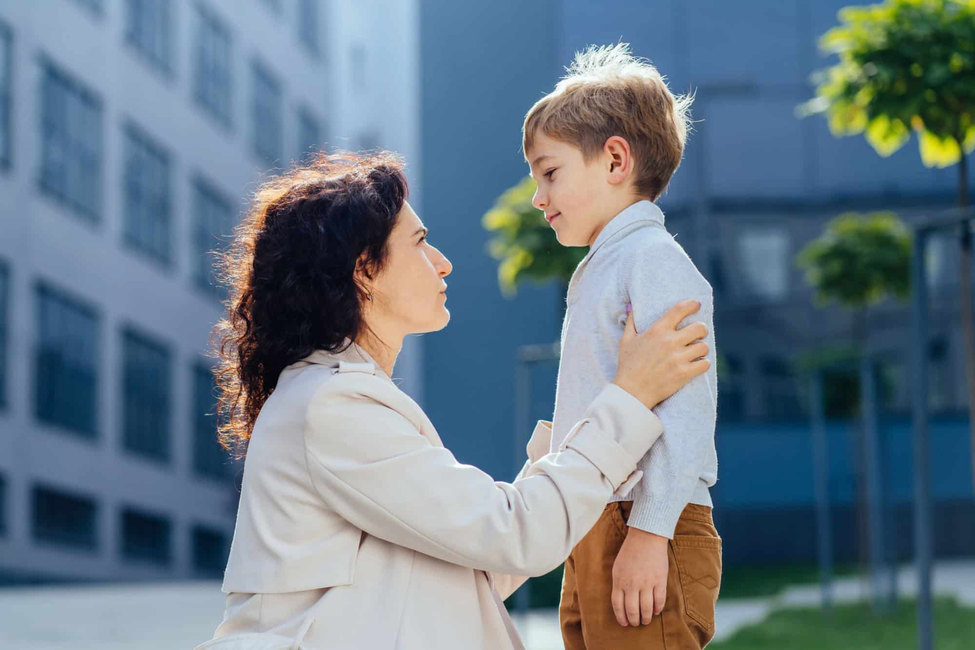 Portrait of mother with brown curly hair who bends to talk to her son, outdoors. Close Up of attractive woman facing young boy. Care, educate, help, raise children, motherhood concept.