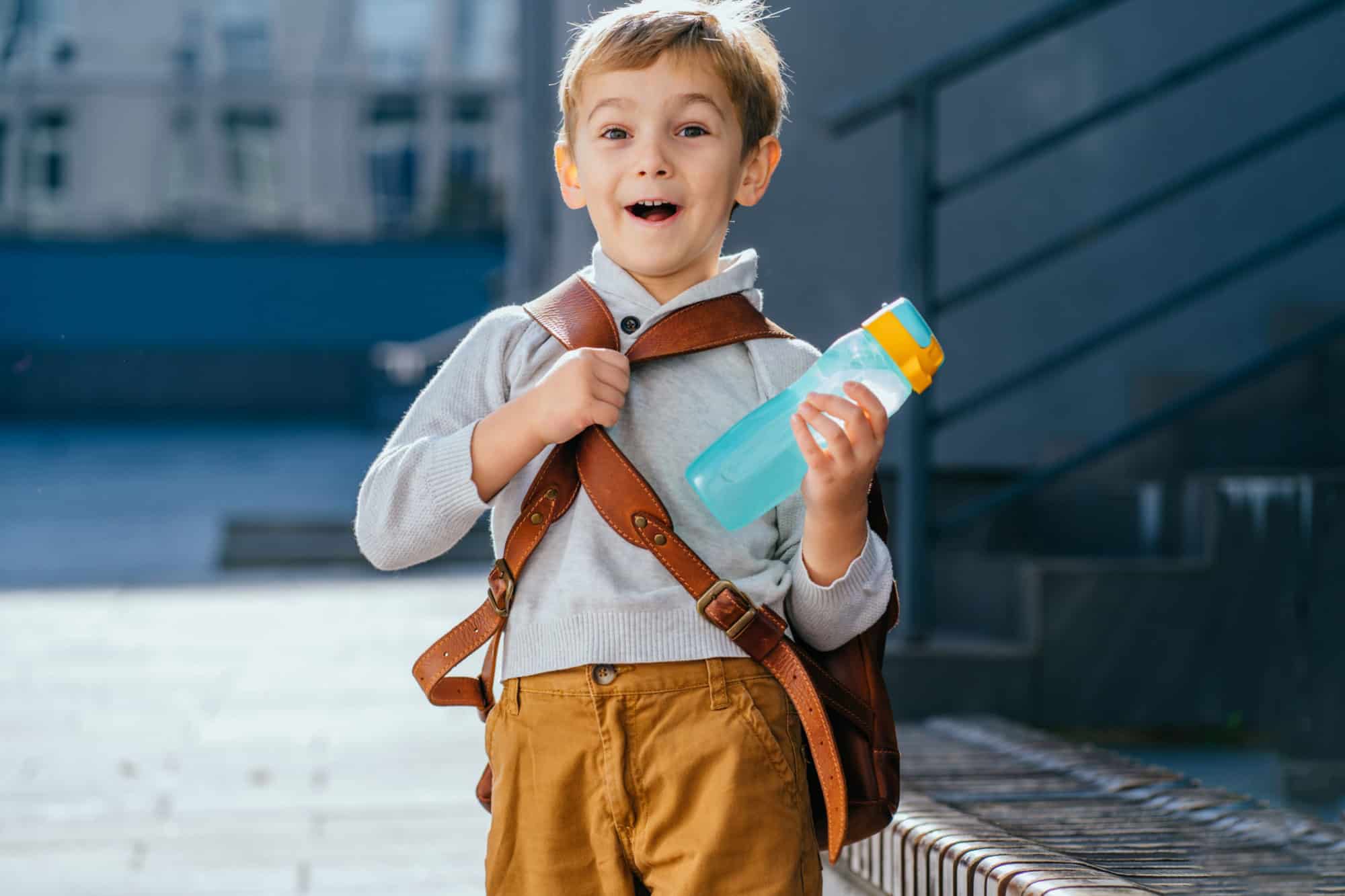 Cute blond little boy going first time to school. Pupil of primary school holding bottle with backpack at sunny day outdoor Walking on School Campus..