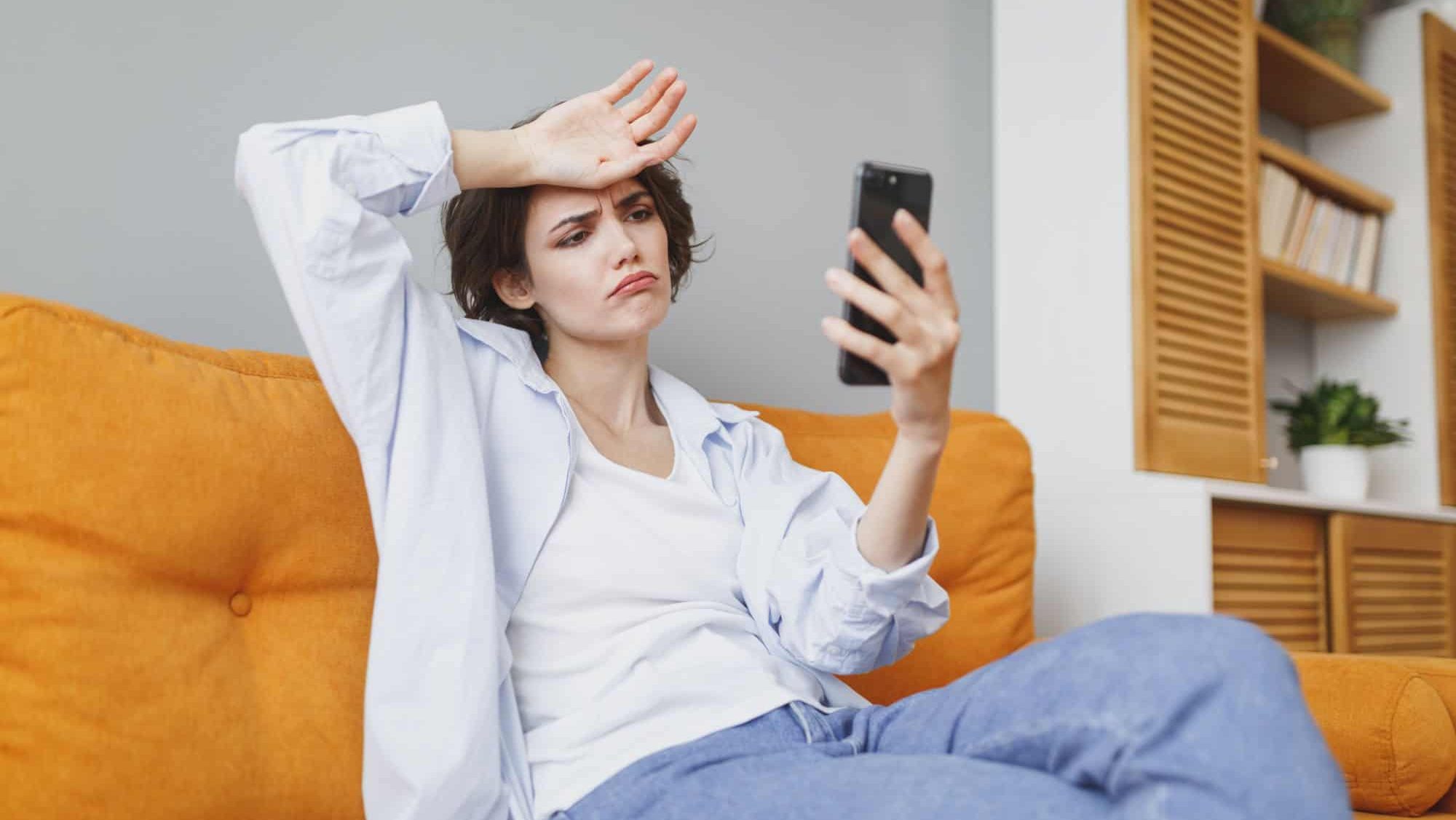 Displeased tired sad woman in casual clothes sit on couch spending time in living room at home. Rest relax good mood leisure concept. Hold using mobile cell phone, taking selfie with and on head