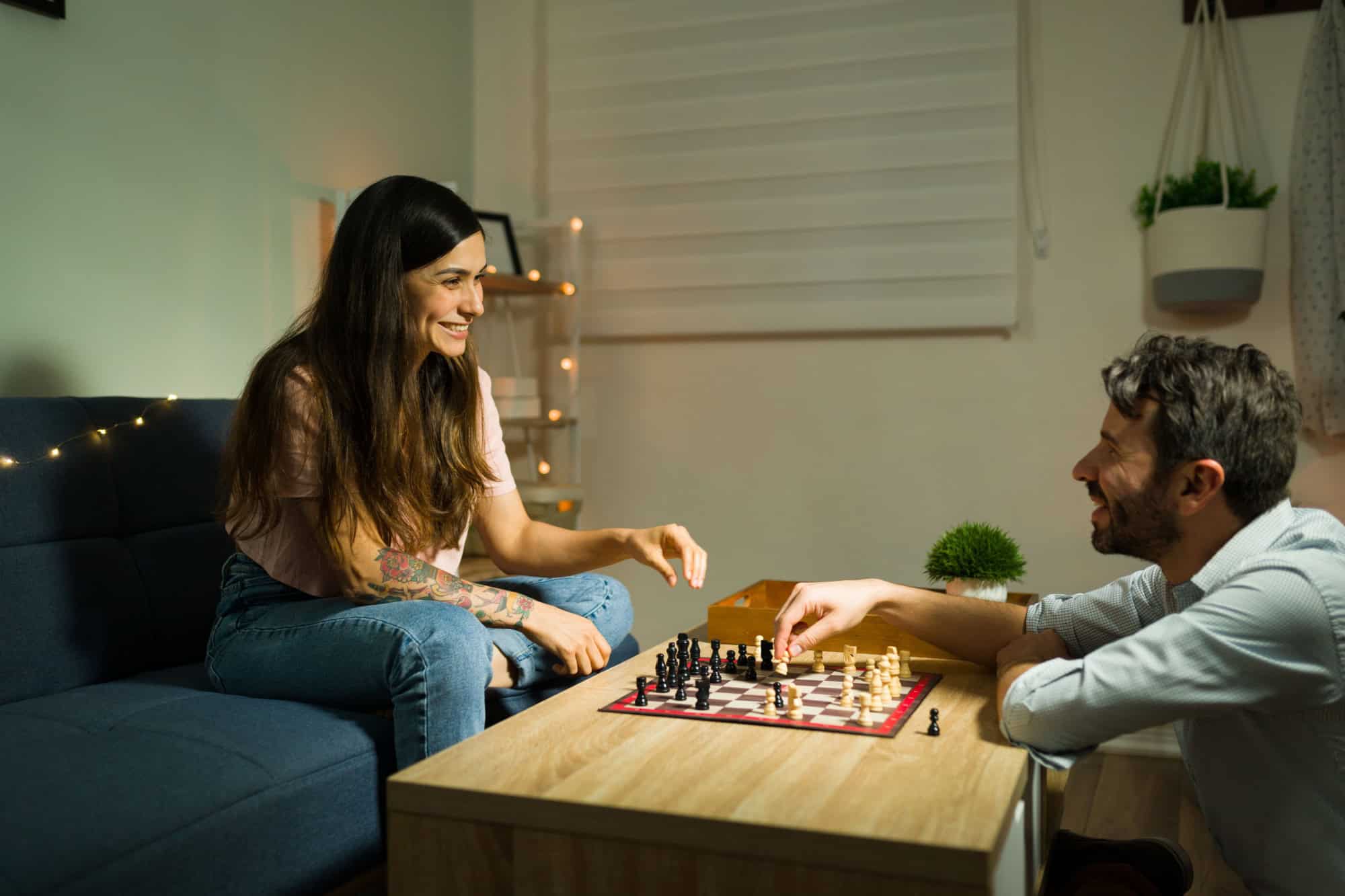 Cheerful young woman laughing while playing chess with her happy partner in the living room coffee table at night