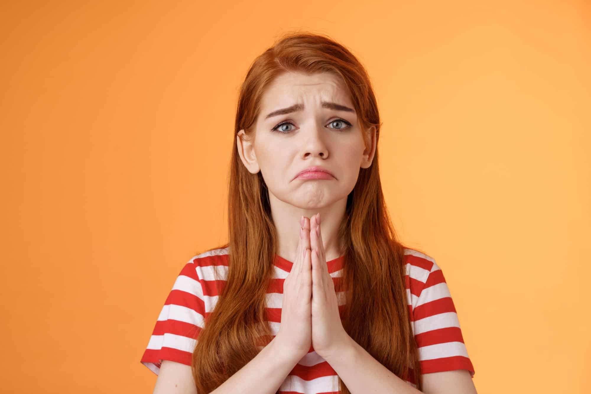 An anxious red-haired woman with a concerned expression and clasped hands, wearing a red and white striped shirt, set against a vibrant orange backdrop.