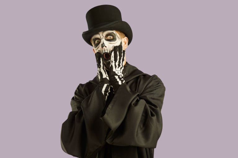 Portrait of shocked man in Halloween make-up and costume grabbing his face in fright. Man in black hat, suit and skull make-up opens his eyes and mouth wide in fear on light lilac background.