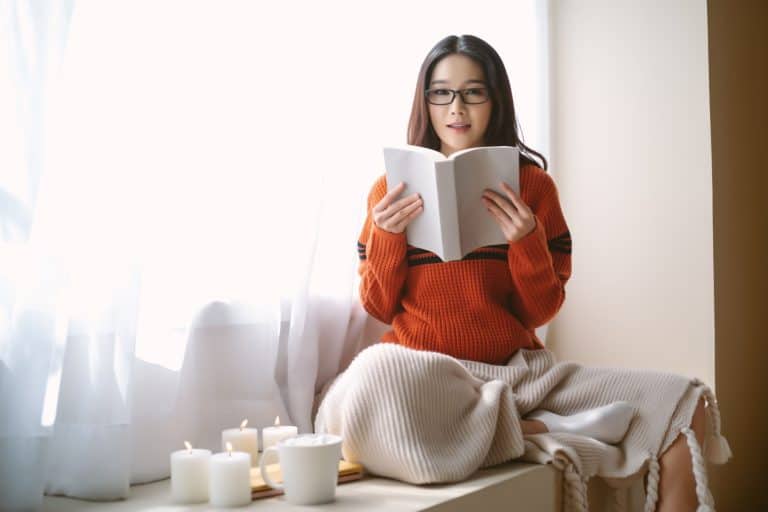 Beautiful young Asian woman sitting in the chair by the window and reading book and looking at camera in bedroom. Happy Influencer Asia girl enjoying free and relax time at home in winter day.