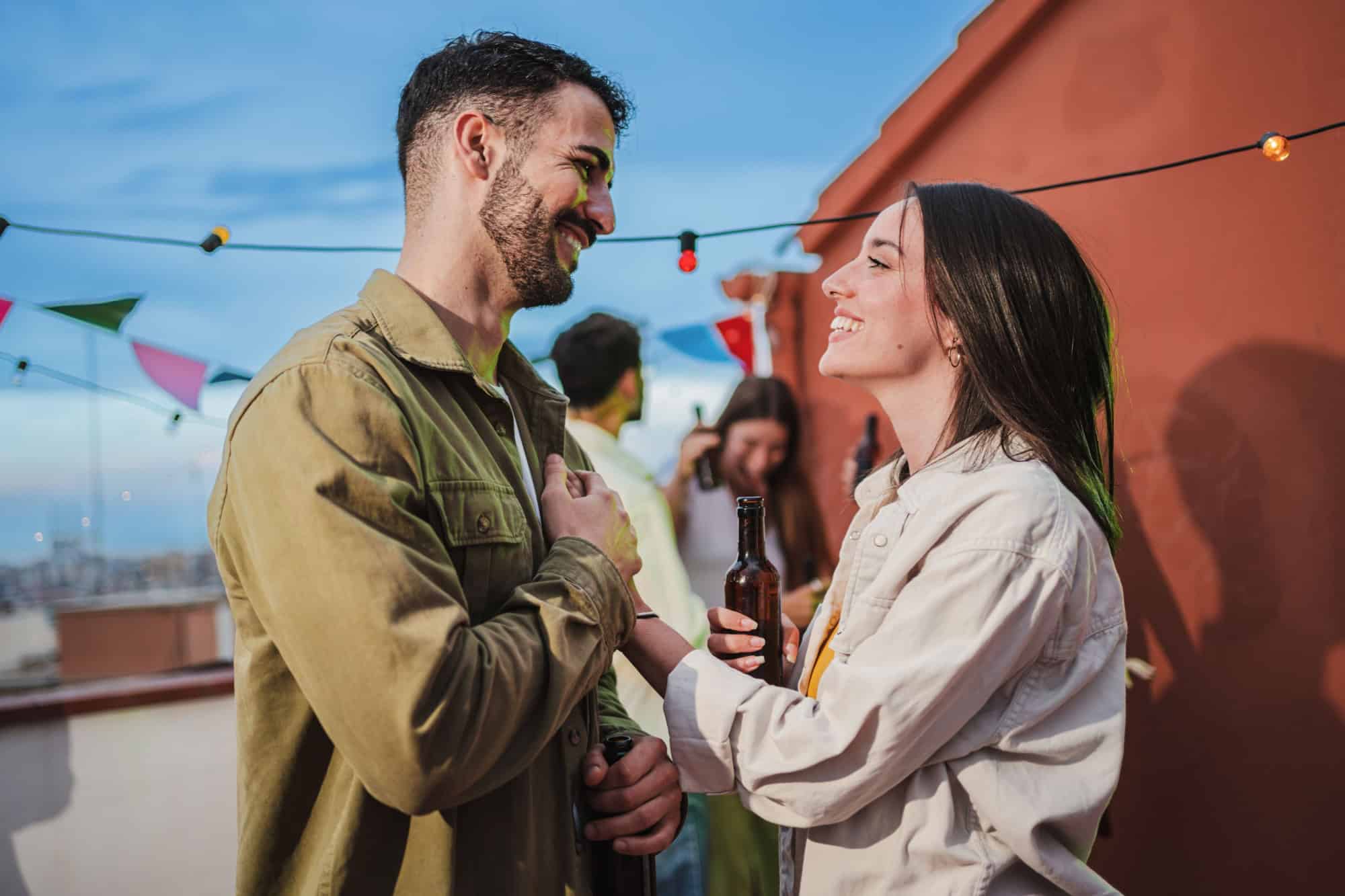 Smiling young couple flirting and having fun at a rooftop party with friends, drinking beer, enjoying and having a pleasant conversation. Handsome guy greeting his joyful girlfriend at celebration