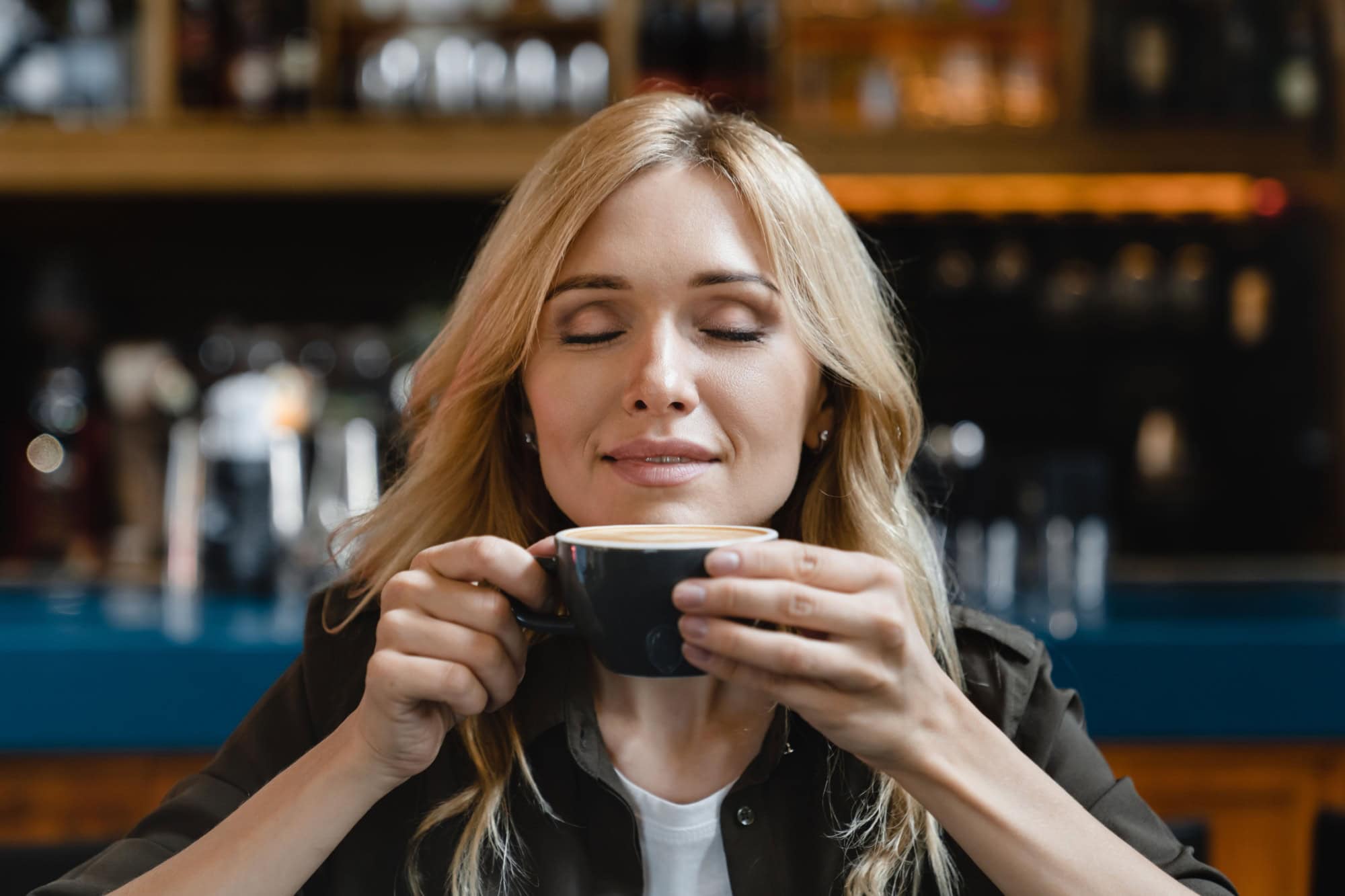 Pretty white young woman student freelancer enjoying aroma of hot beverage coffee tea smelling it while having breakfast workbreak brunch in restaurant cafe.