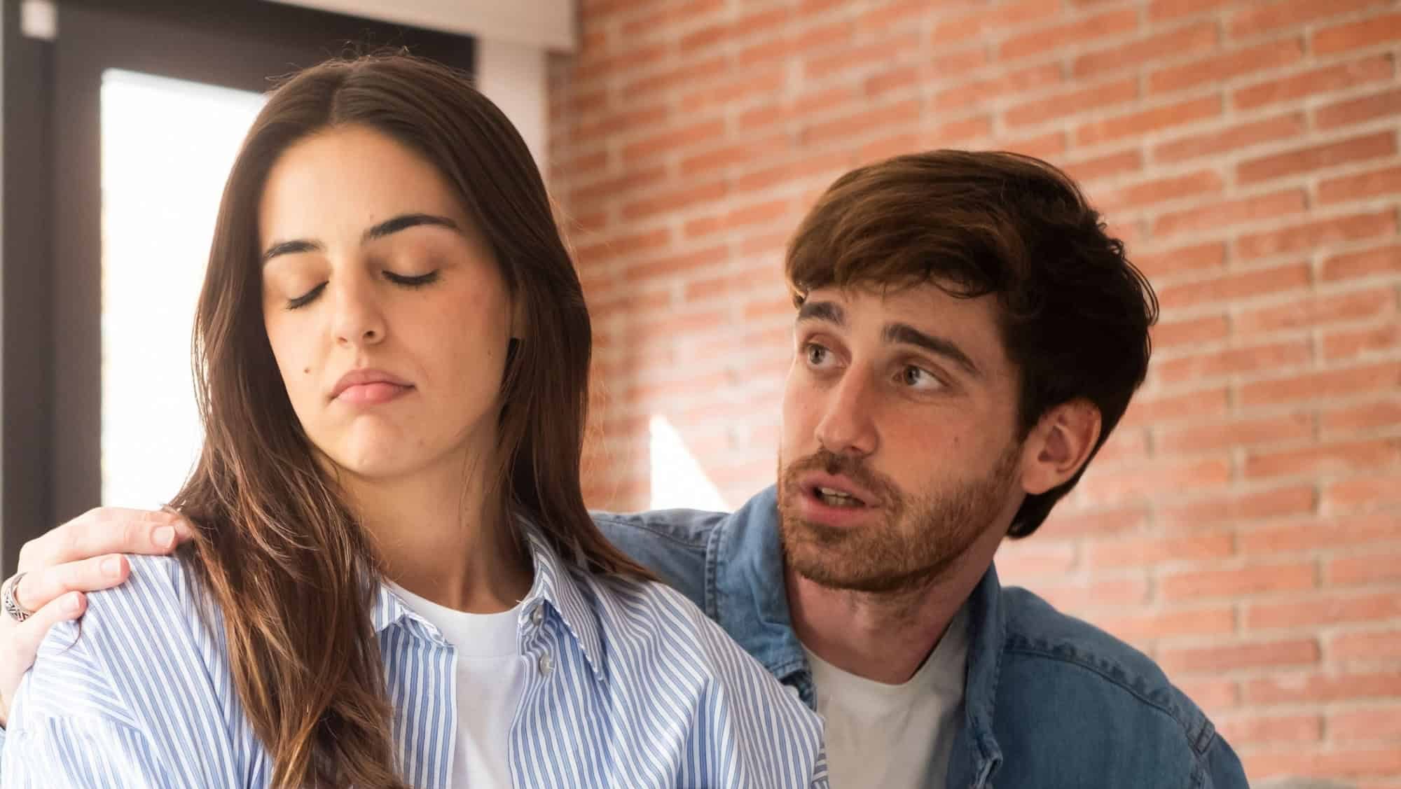 A young couple faces emotional turmoil at home during a painful breakup discussion. The offended woman ignores her sad boyfriend as he tries to justify himself, reflecting the tension of infidelity
