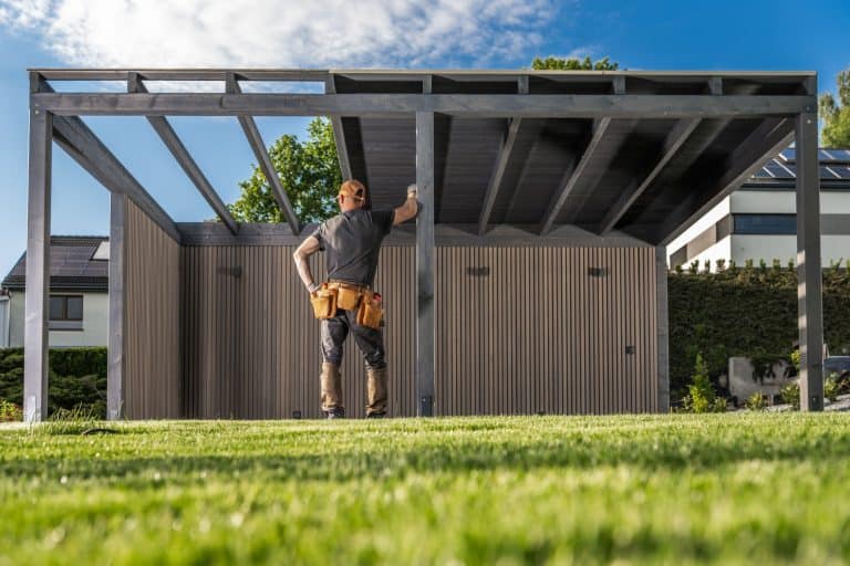 A construction worker stands in a grassy yard, evaluating a newly built outdoor structure with a contemporary design on a clear day.