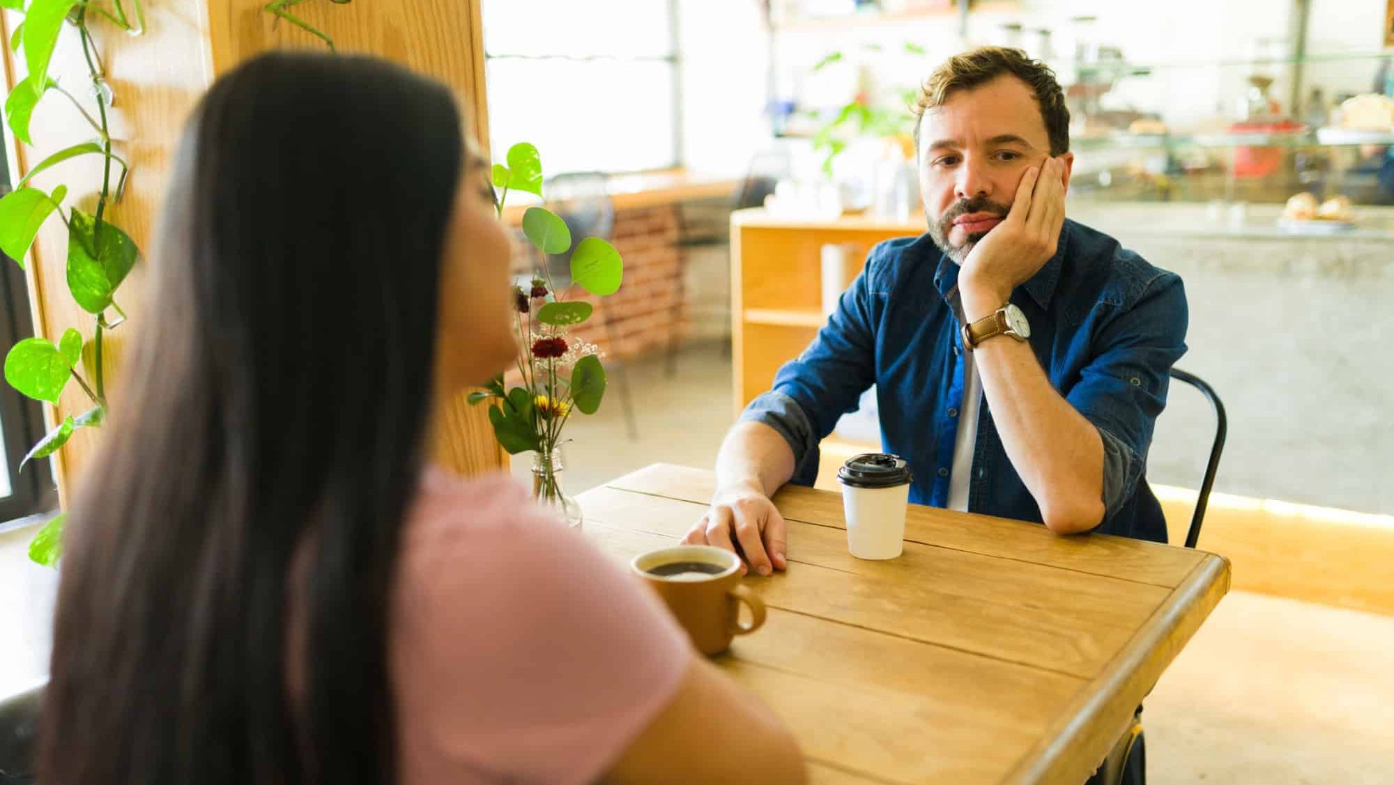 Bored man ignoring his girlfriend while sipping coffee at the cafe, highlighting communication issues in their troubled relationship