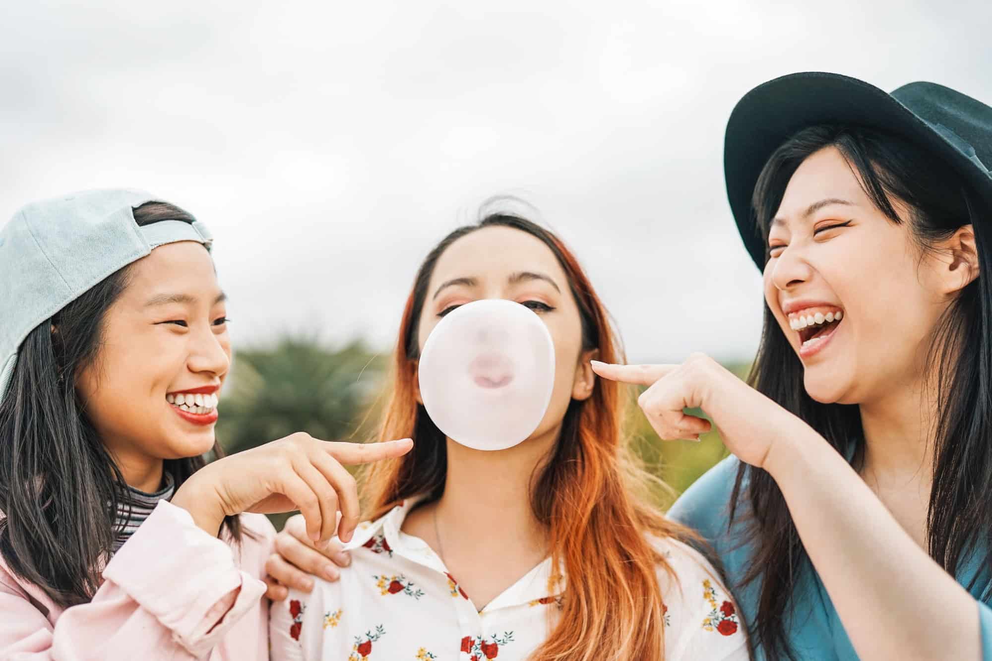 Happy Asian friends having fun chewing bubble gum outdoor - Young people playing and laughing together