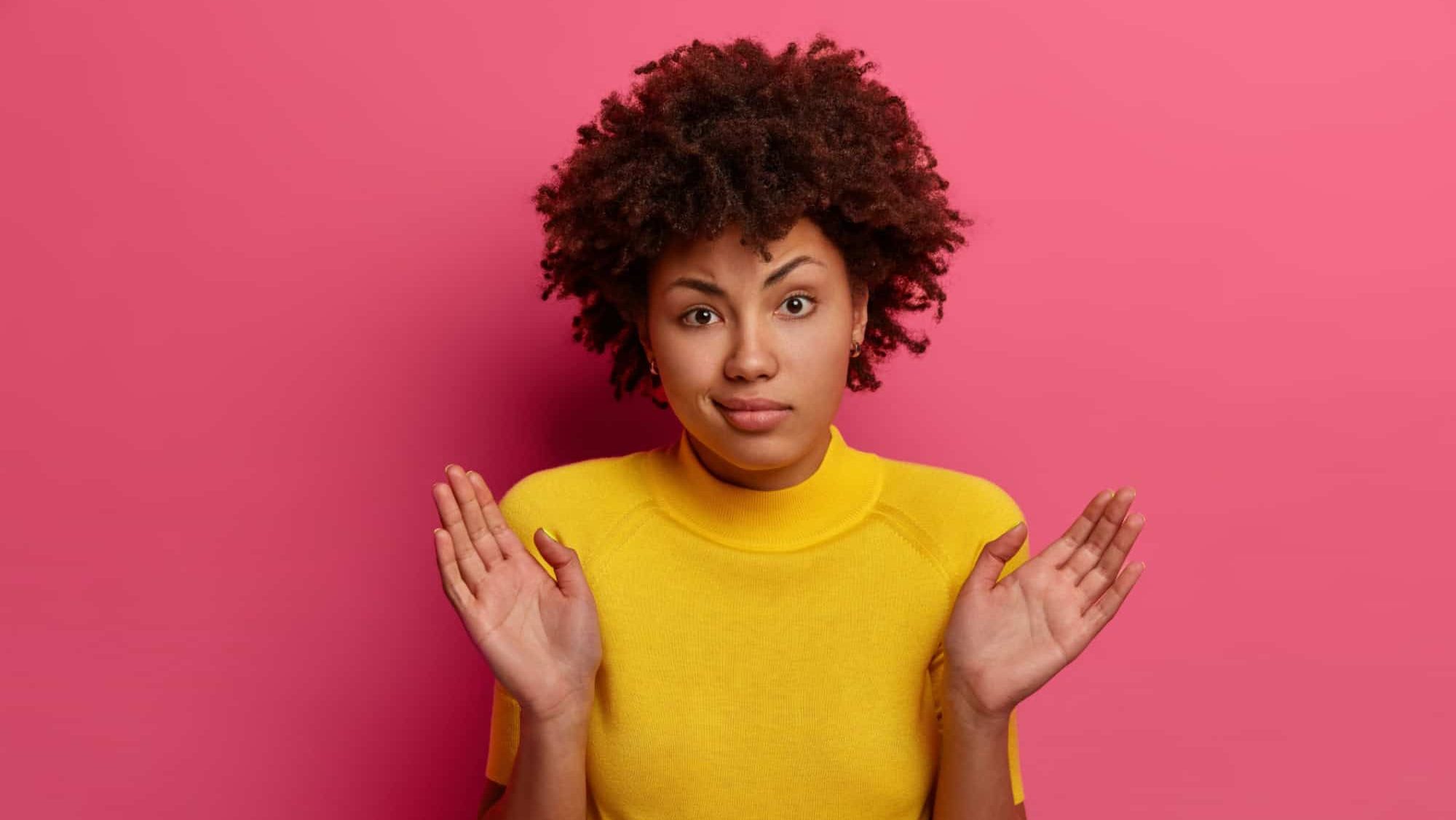 Perplexed Afro American woman raises palms with hesitation, cant help, stands confused and indecisive, says its not my fault, wears yellow outfit, poses over pink background, shrugs shoulders