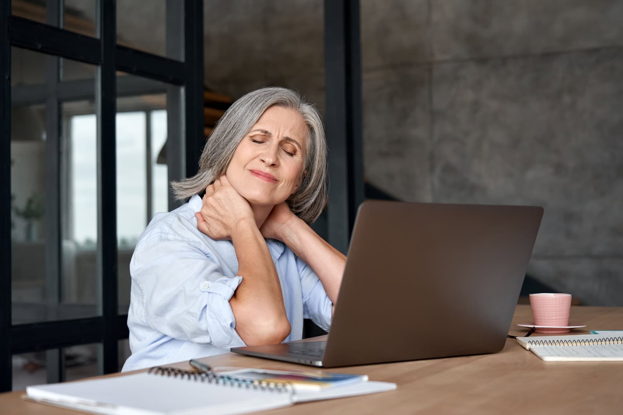 Tired stressed old mature business woman suffering from fibromyalgia neck pain working in office sitting at table. Overworked senior middle aged lady massaging neck feeling hurt pain from sedentary job