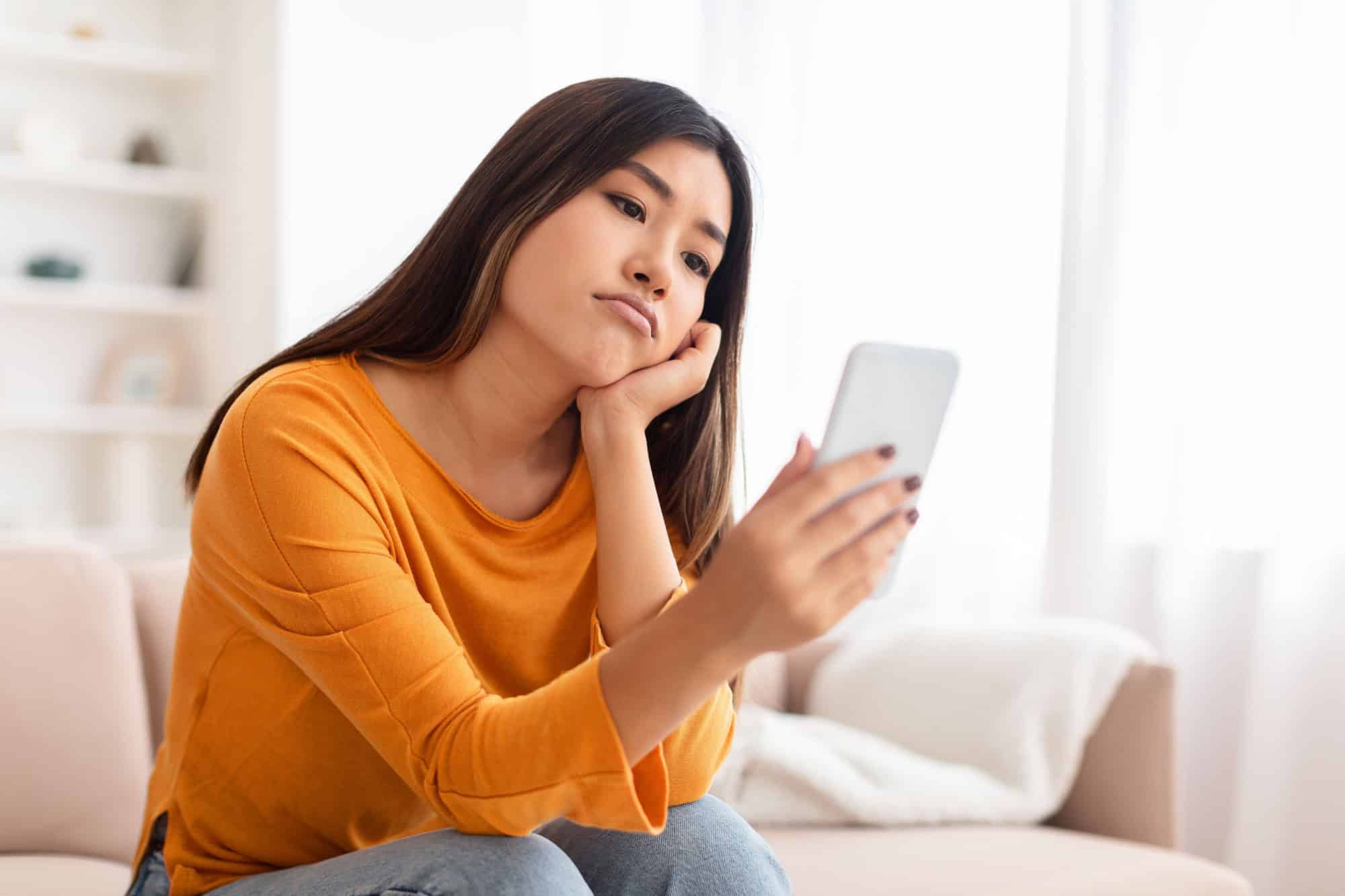 Smartphone Addiction And Depression. Frustrated bored asian young lady using mobile phone, checking social media, reading bad news sitting on the couch. Upset woman waiting for his call, copy space