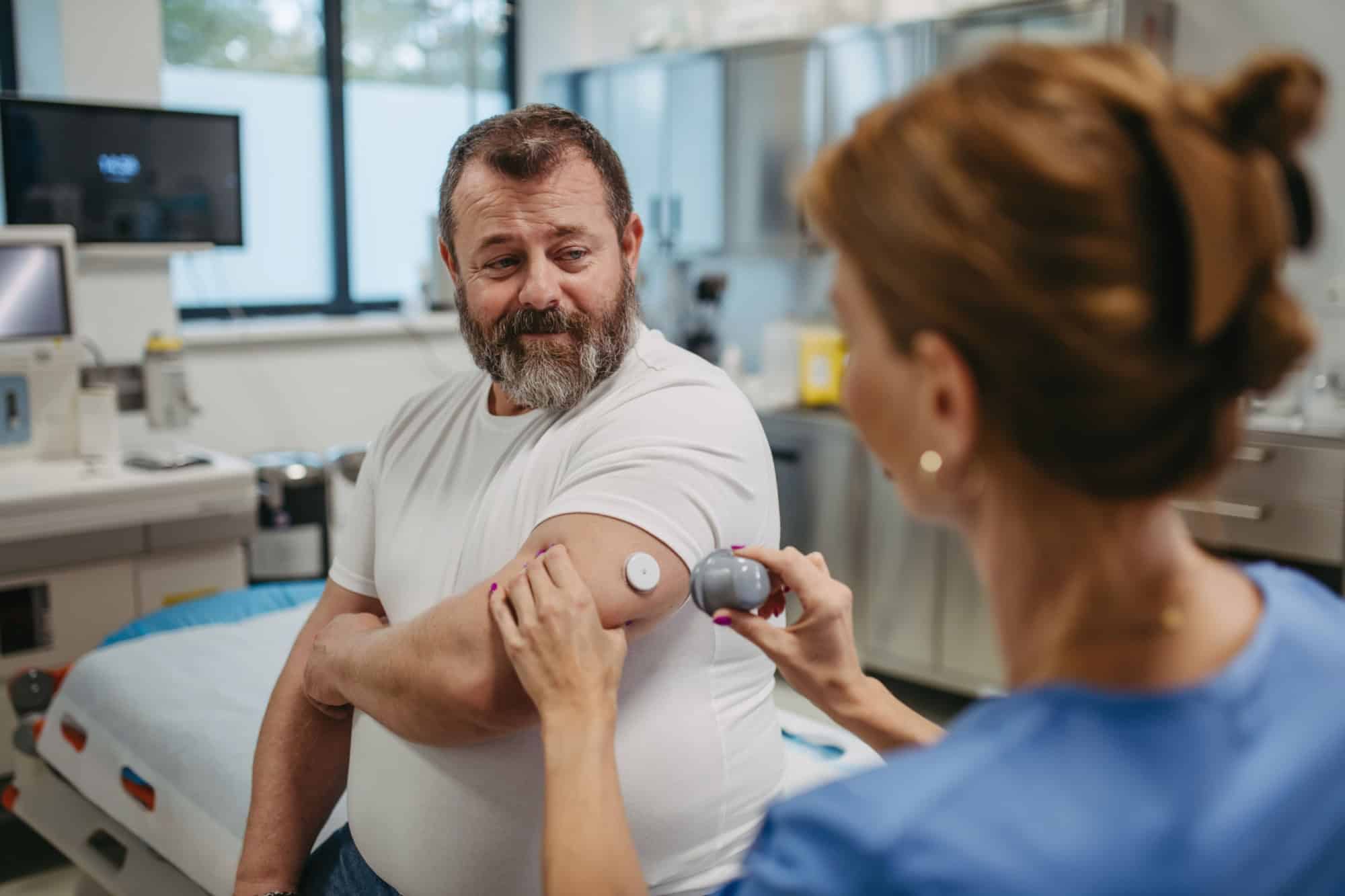 Doctor applying a continuous glucose monitor sensor on patient's arm. Obese, overweight man is at risk of developing type 2 diabetes. Concept of health risks of overweight and obesity.