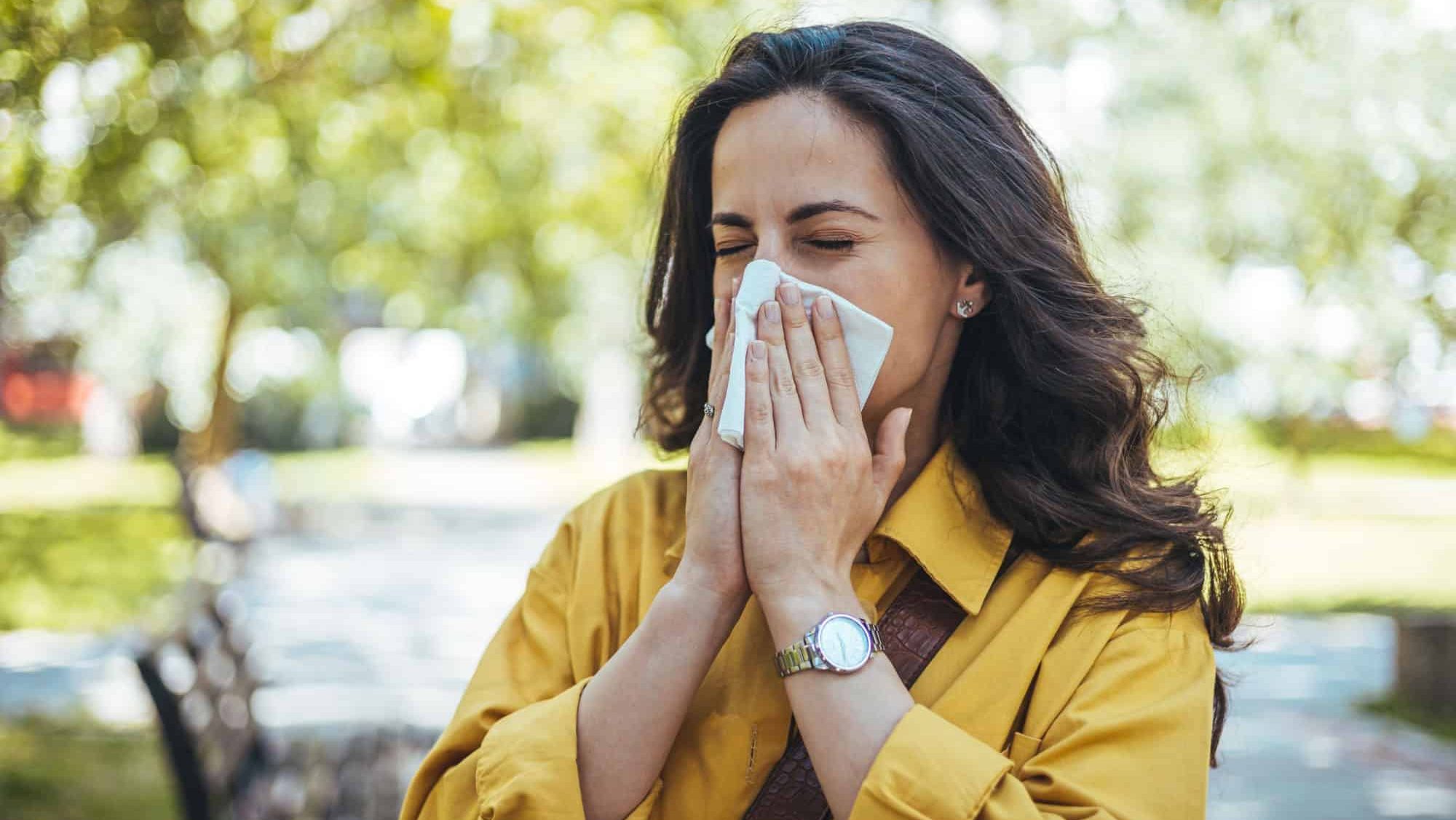 Shot of an attractive young woman feeling ill and blowing her nose with a tissue outdoors. Woman has sneezing. Young woman is having flu and she is sneezing. Sickness, seasonal virus problem concept