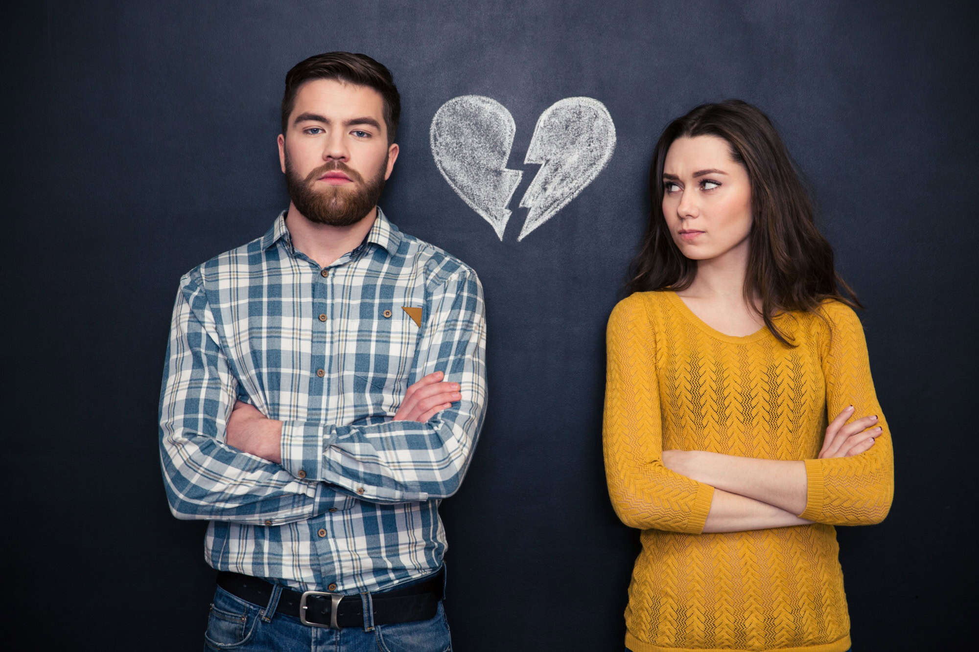 Portrait of young couple after argument standing separately with hands folded over blackboard background
