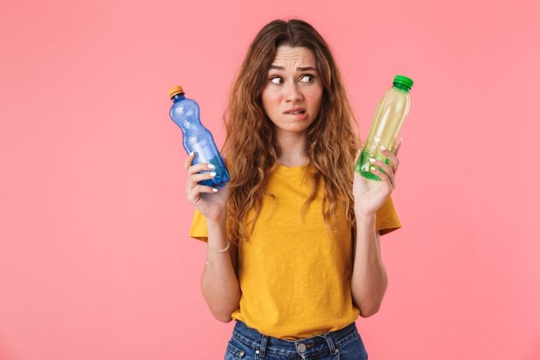 Photo of confused caucasian woman in basic t-shirt holding plastic bottles and looking aside isolated over pink background