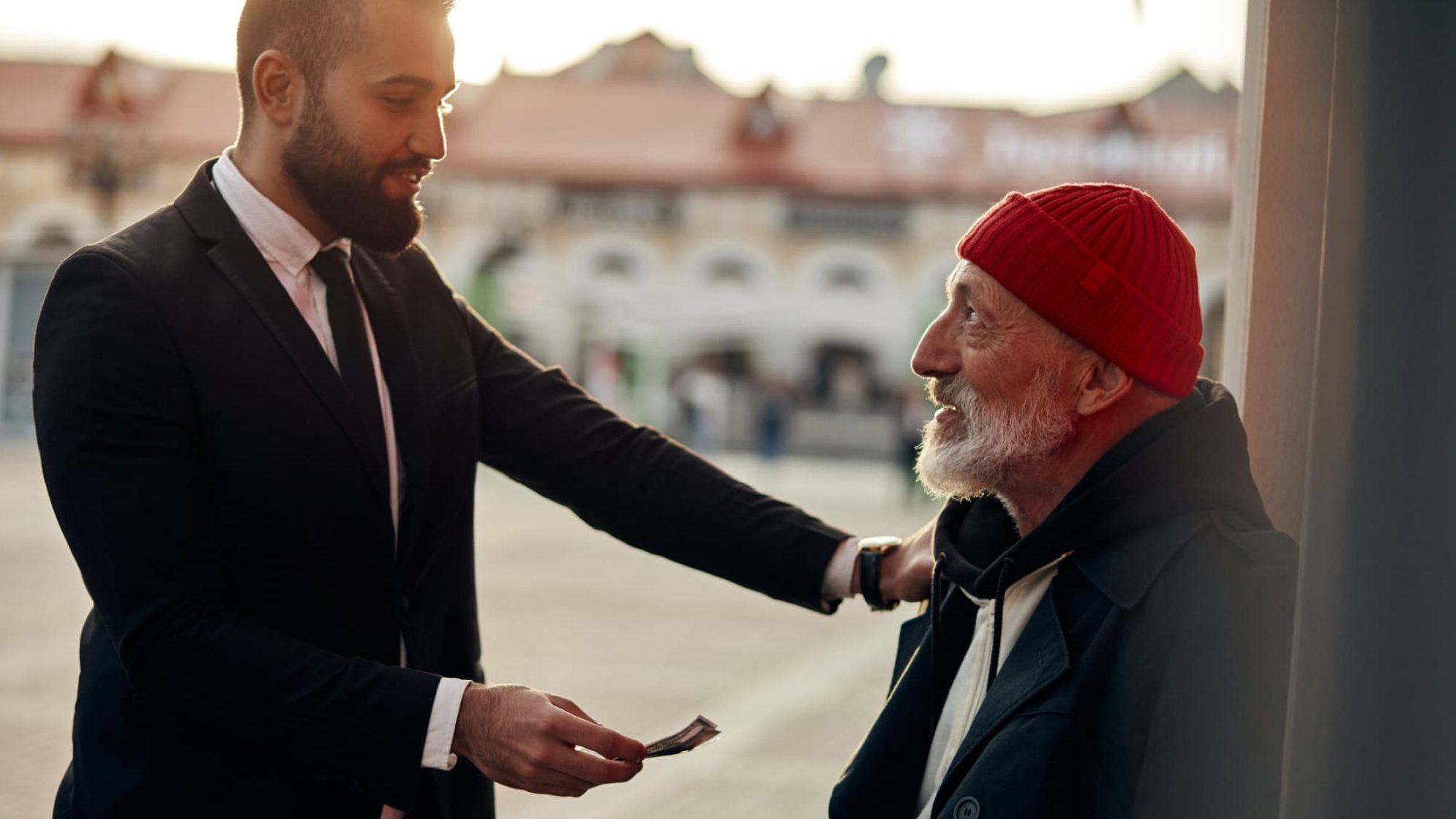 Businessman in tuxedo help to vagrant man sitting on street with rusty money jar. Rich man put one hand on shoulder of homeless person and give one dollar. Kind people, compassion, sympathy