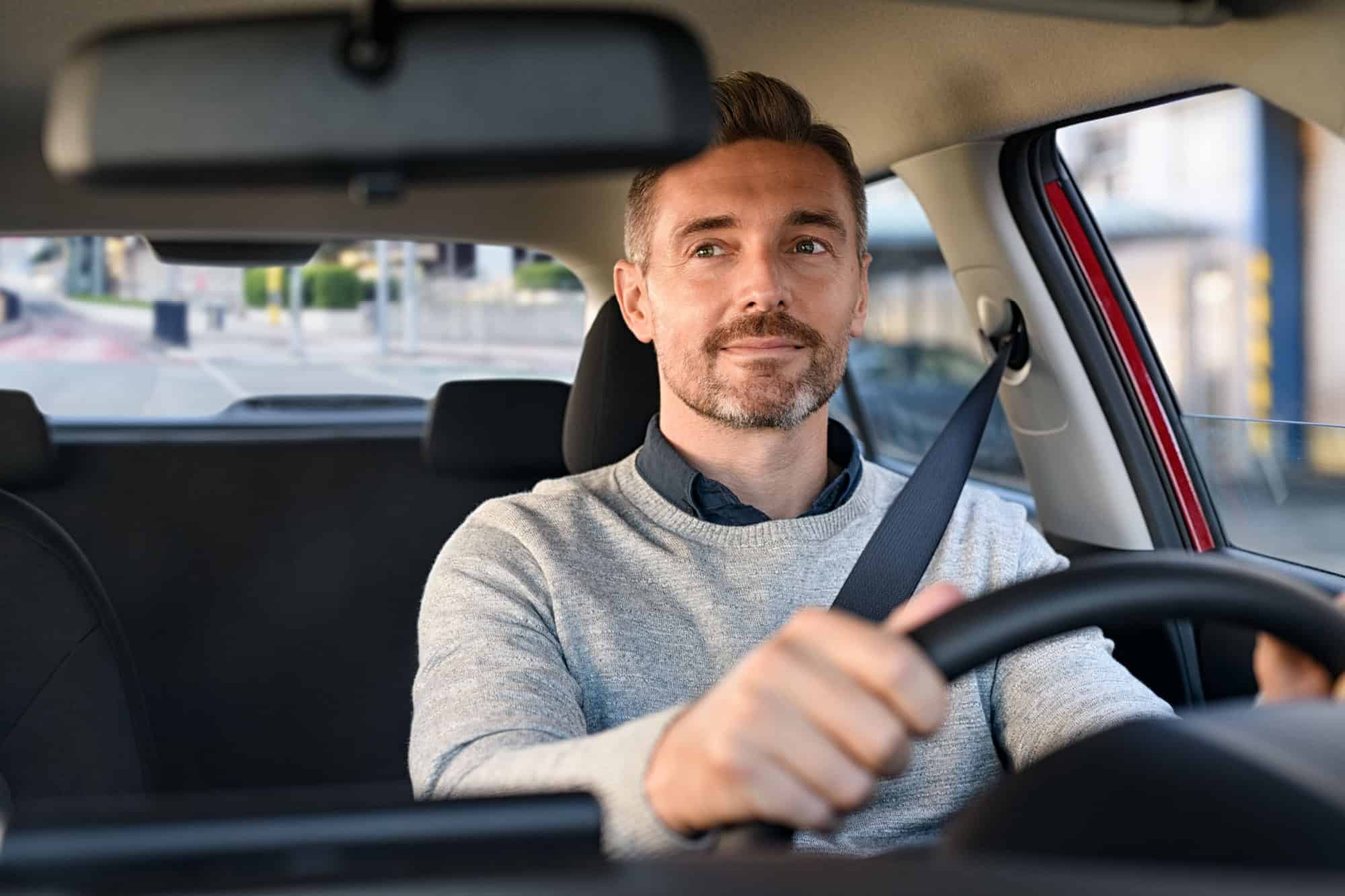 Mid adult man smiling while driving car and looking at mirror for reverse. Happy man feeling comfortable sitting on driver seat in his new car. Smiling mature businessman with seat belt on driving.