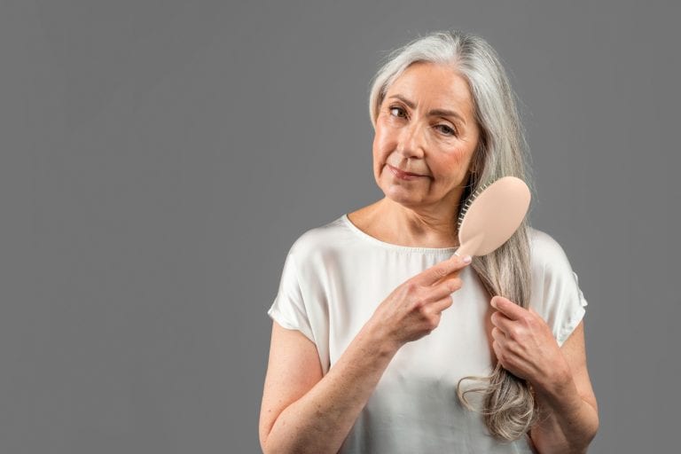 Serious senior caucasian female combing her gray hair with comb, isolated on gray background, studio, copy space. Beauty care at home daily procedure, treatments and problems with hair, ad and offer