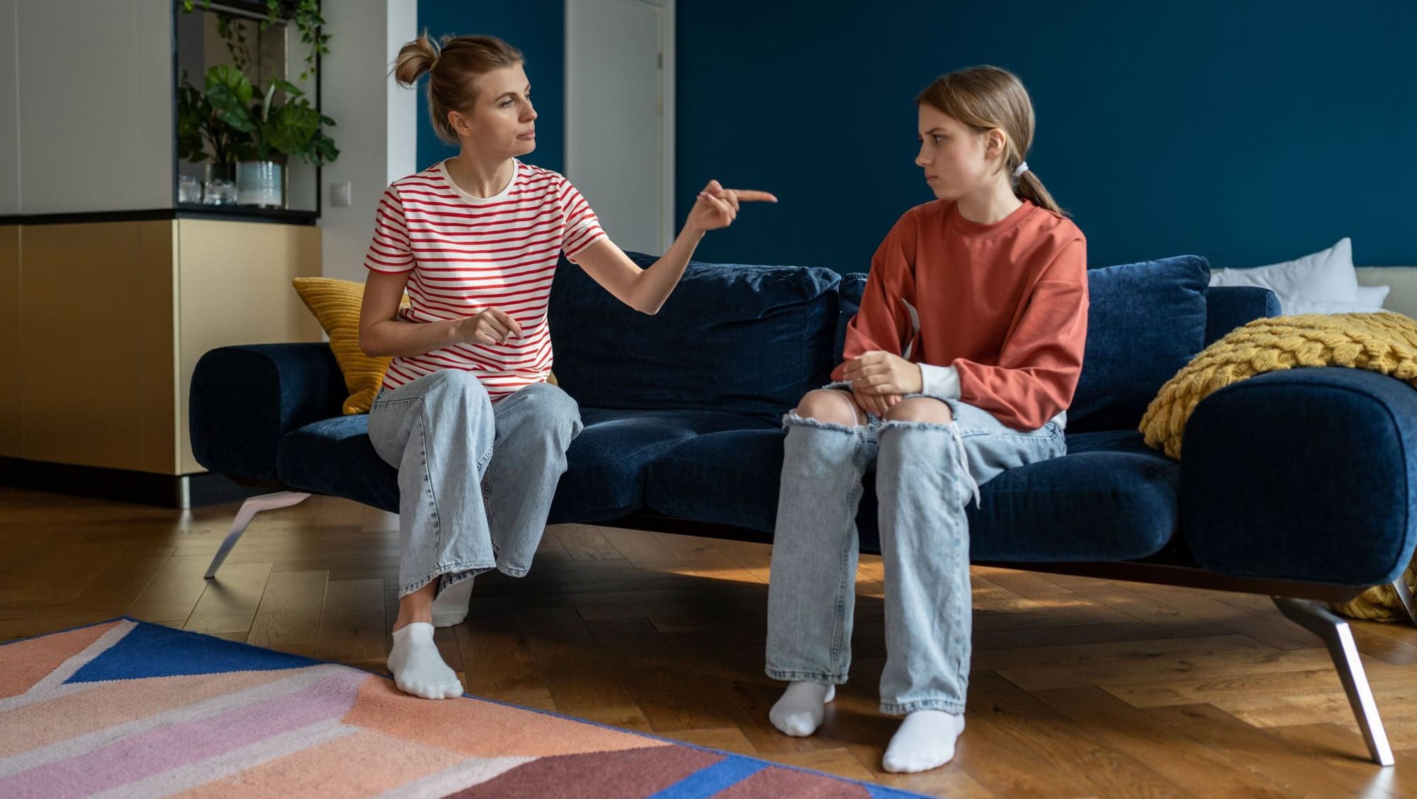 Parent-teen conflict. Strict mother scolding lecturing upset teenage girl daughter at home, pointing finger. Mom disciplining teenager while sitting together on sofa. Parenting adolescent