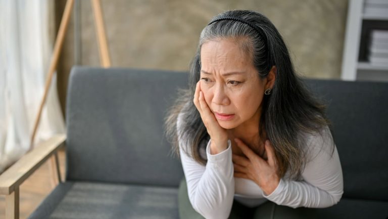 Shocked and concerned Asian mature woman look in the distance, thinking and worried about something while sitting on sofa in her living rom, panic attack, anxiety, depressed.