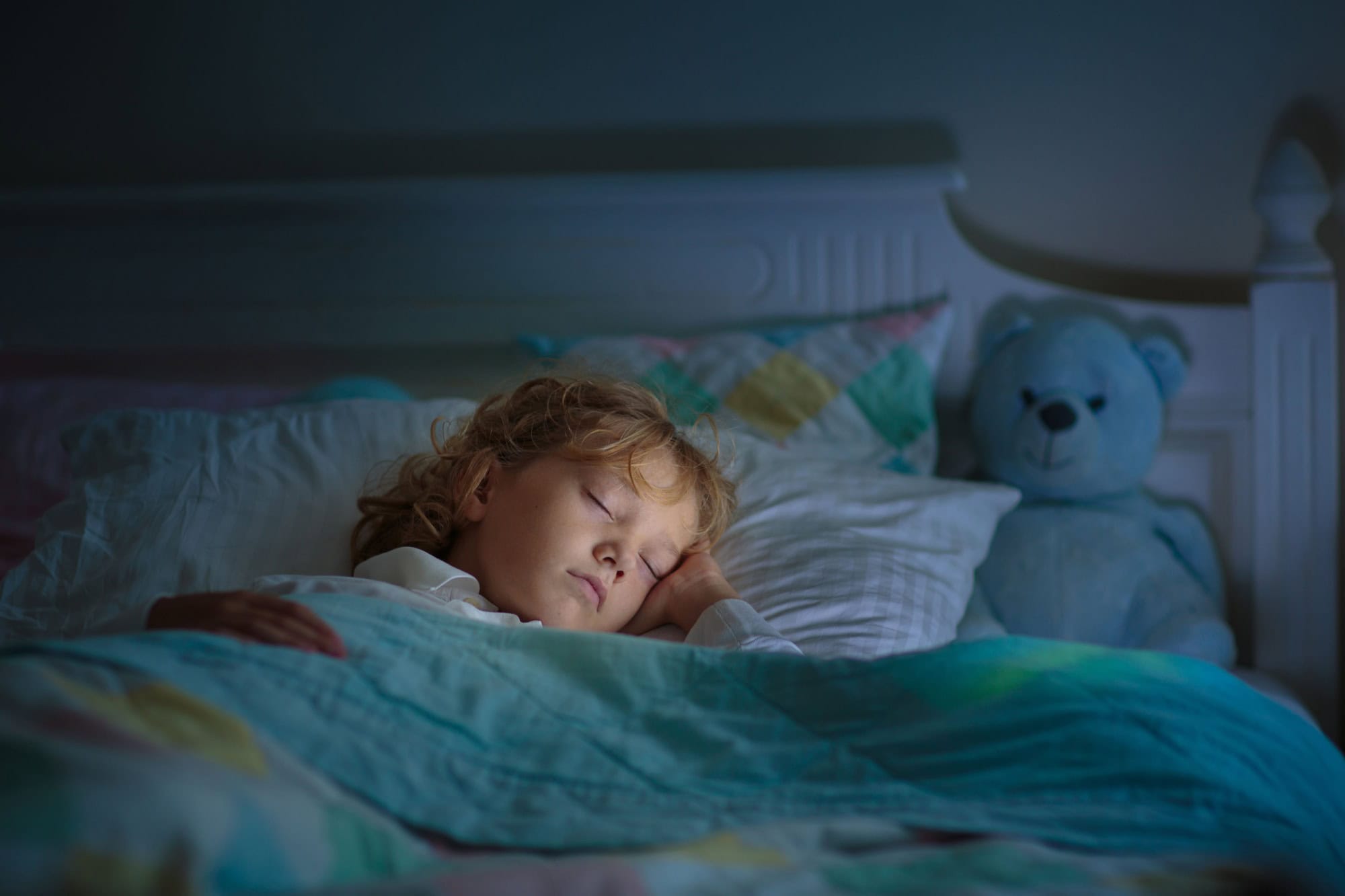 Child sleeping in dark bedroom. Little boy napping. Healthy night rest. Kids room with night light. Toddler with teddy bear. Bedding for children.