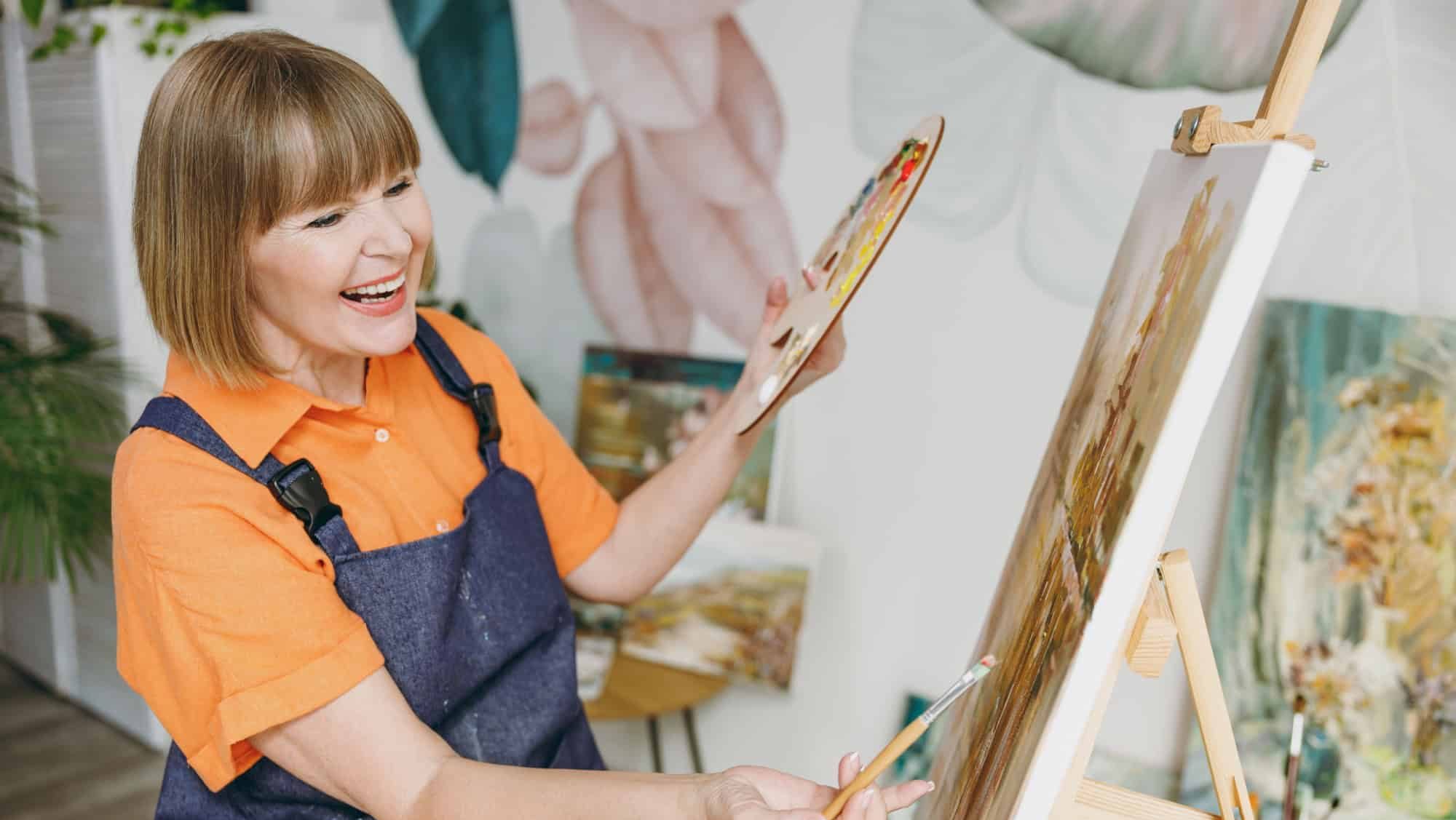 Side view elderly smiling artist woman 50 years old wear casual clothes sitting near easel with painting artwork paint hold palette spend free spare time in living room indoor. Leisure hobby concept