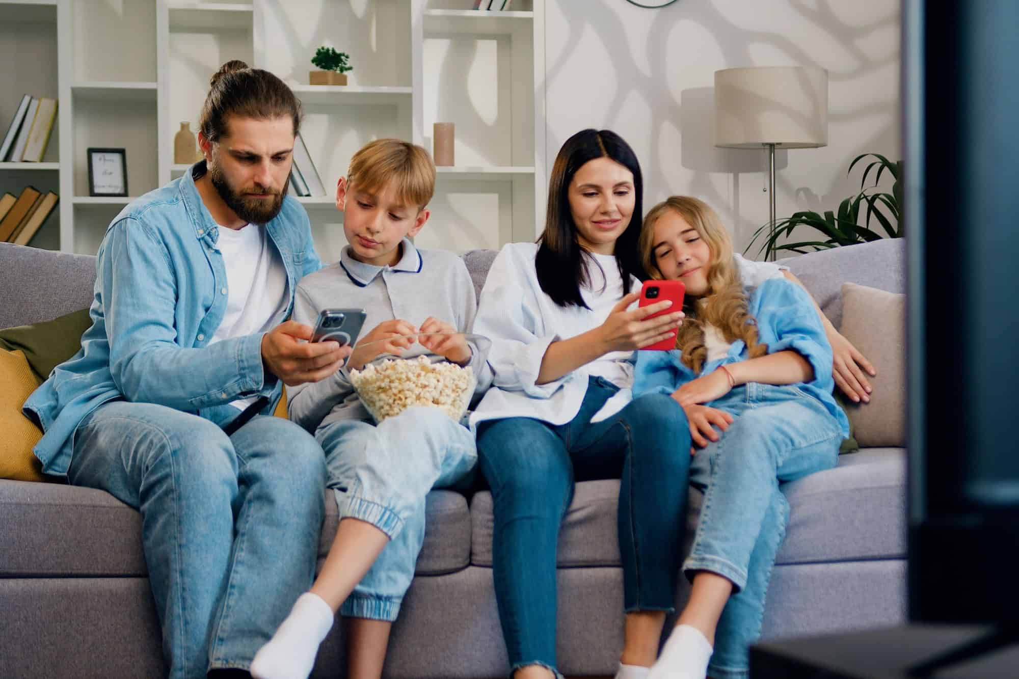 Attractive sad family sitting together on sofa couch at home and everybody using smartphone to read messages or chatting with friends.