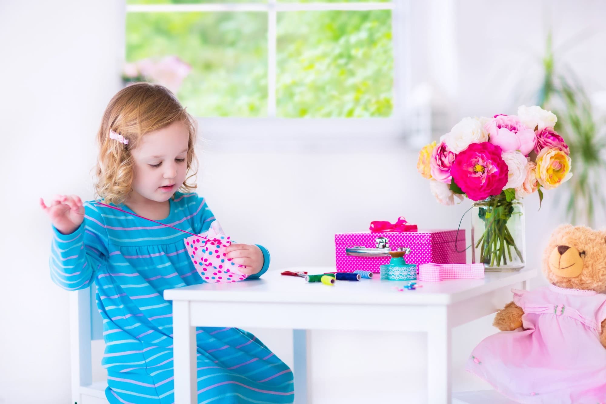 Cute creative little girl sewing a dress for her teddy bear doll, playing with needles and ribbons in a white sunny room at home or preschool