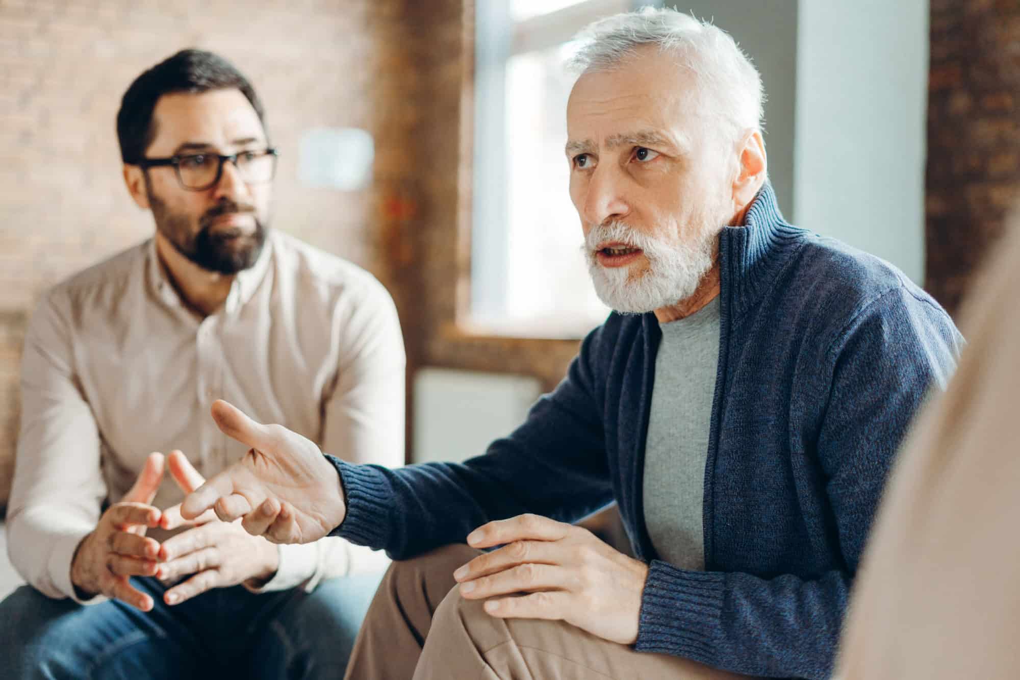Senior man gesturing while explaining something during group therapy session, sharing his problems and trying to find solutions with therapist and other patients. Mental health care concept