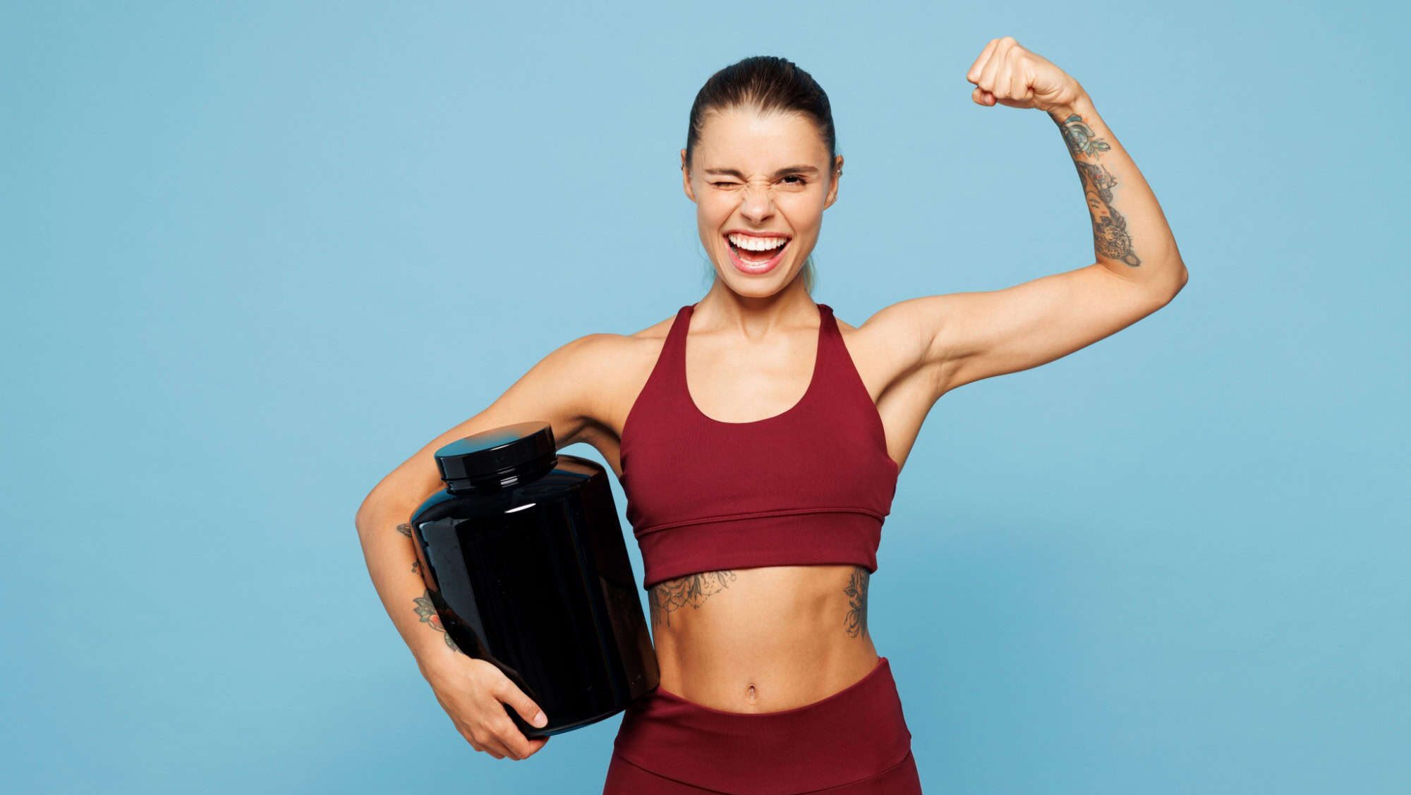 Young fitness trainer instructor sporty woman wear red sportswear training in home gym hold protein supplements bottle show muscles wink isolated on plain blue background Workout sport fit abs concept